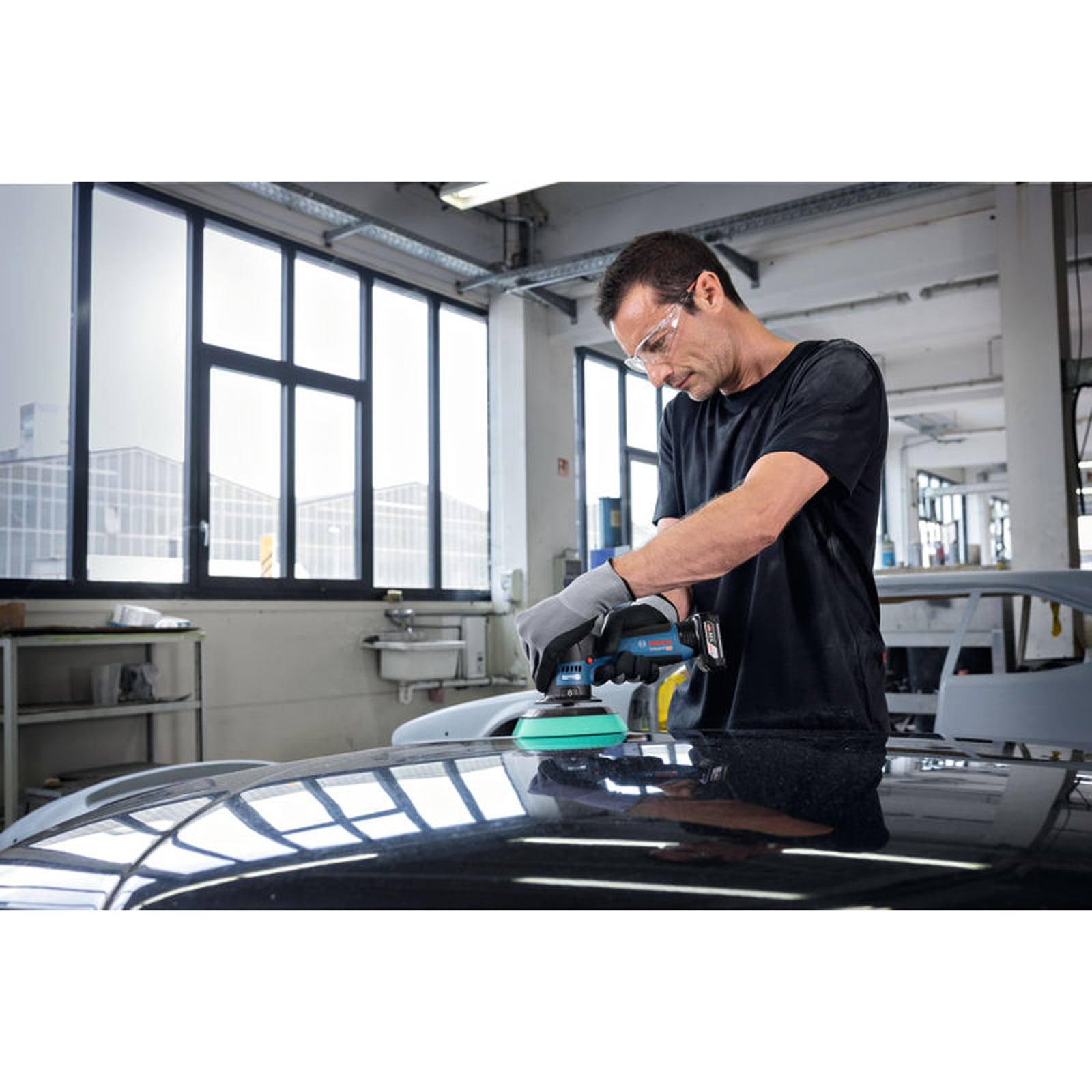 Ein Handwerker poliert mit einer blauen Poliermaschine die schwarze Motorhaube eines Autos in einer Werkstatt.