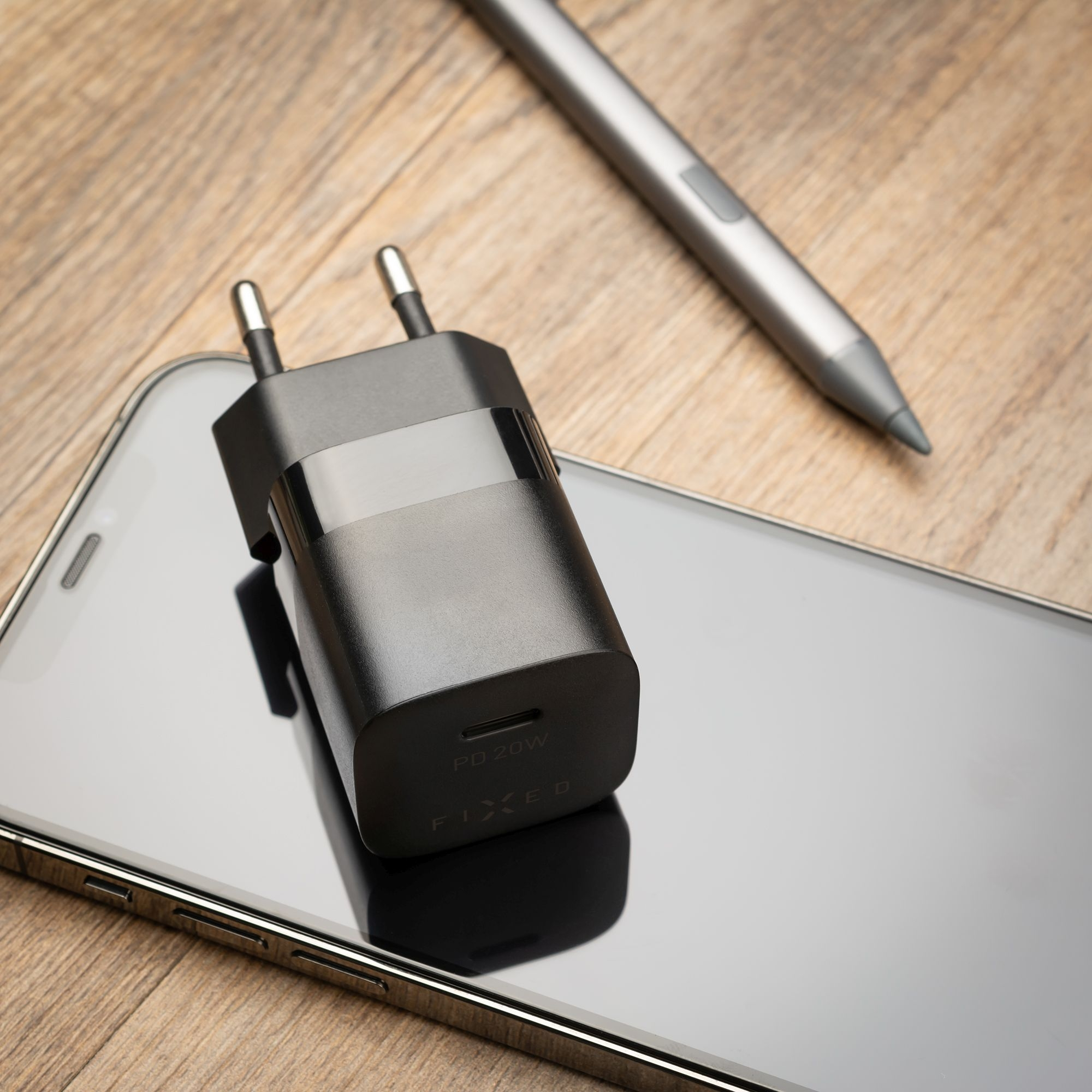 Ein schwarzes Fixed PD 20W USB-Ladegerät liegt auf einem Smartphone neben einem Stift auf Holztisch.