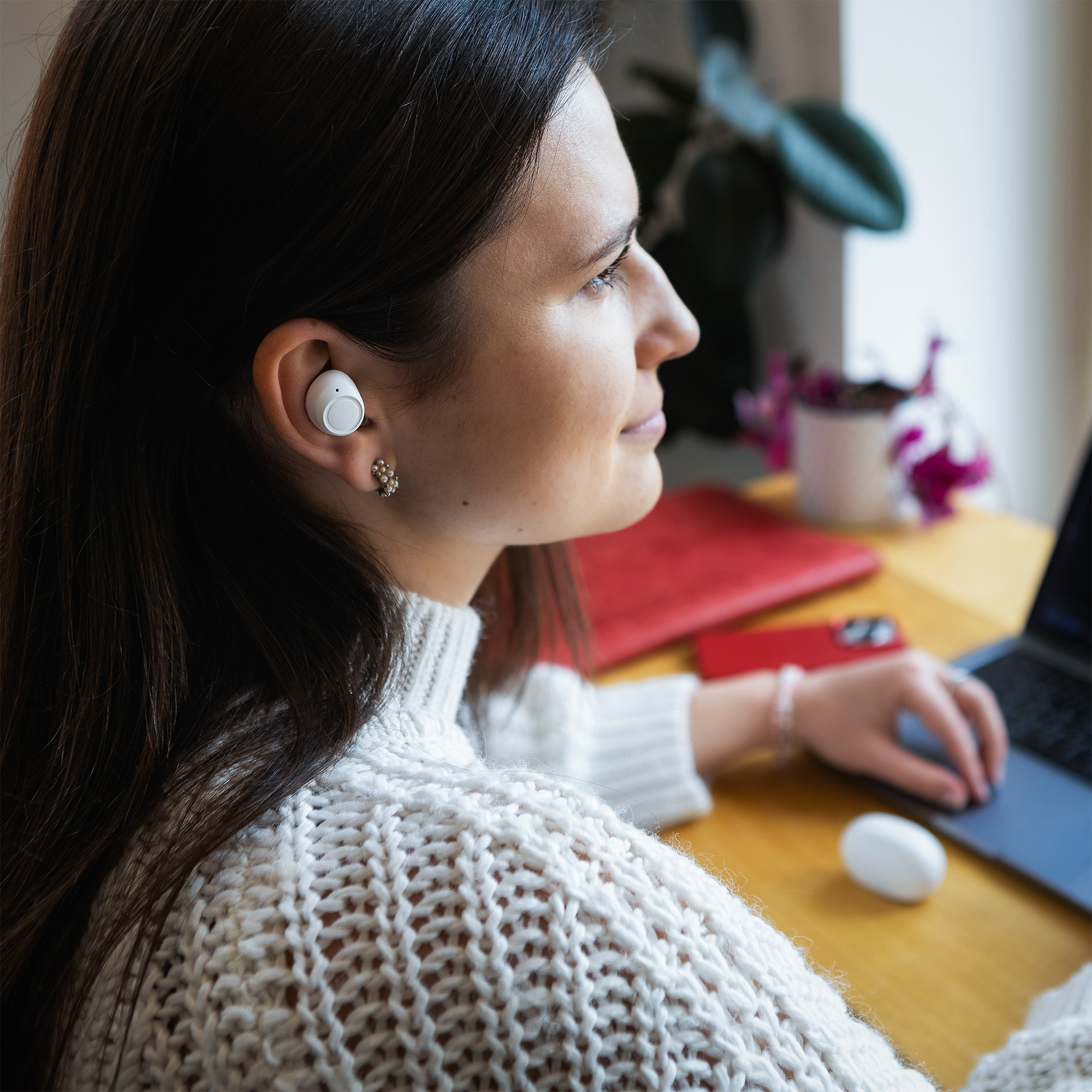 Eine brünette Frau trägt weiße In-Ear-Kopfhörer und arbeitet an einem Holztisch an einem Laptop.