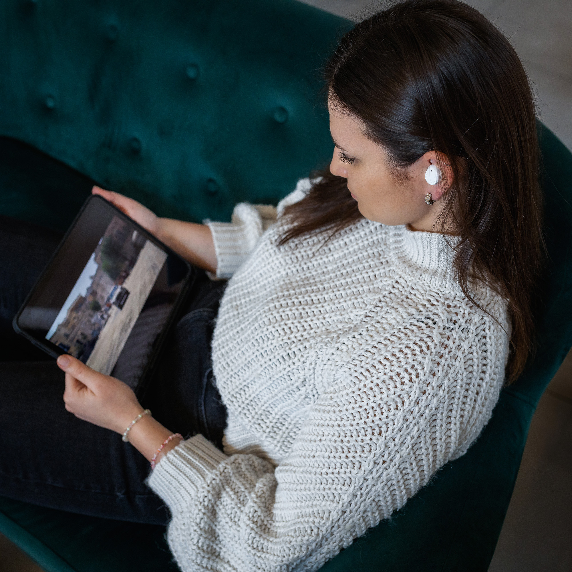 Eine Frau in einem weißen Pullover auf einer grünen Couch hält ein Tablet und trägt weiße Ohrhörer.