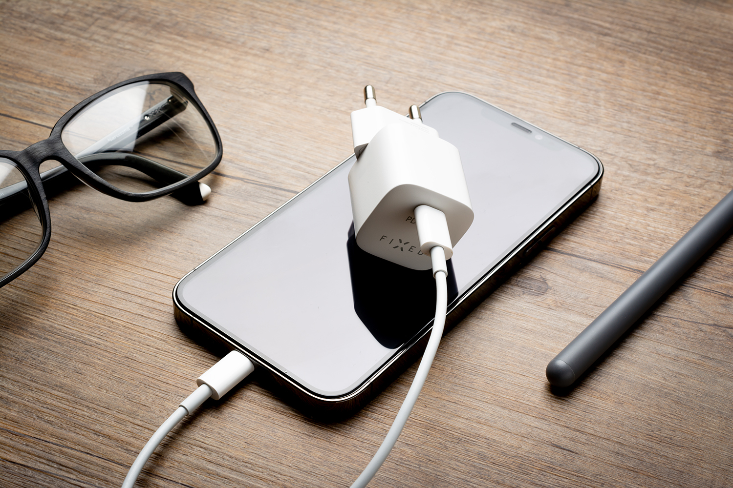 Ein schwarzes Smartphone mit weißem Ladekabel und Adapter auf Holztisch, daneben Brille und Stift.