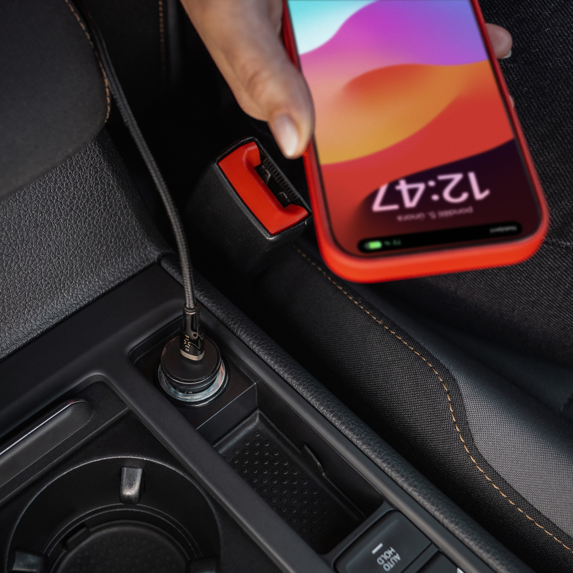 Eine Hand hält ein Handy mit roter Hülle über die Mittelkonsole eines Autos, Ladekabel eingesteckt.