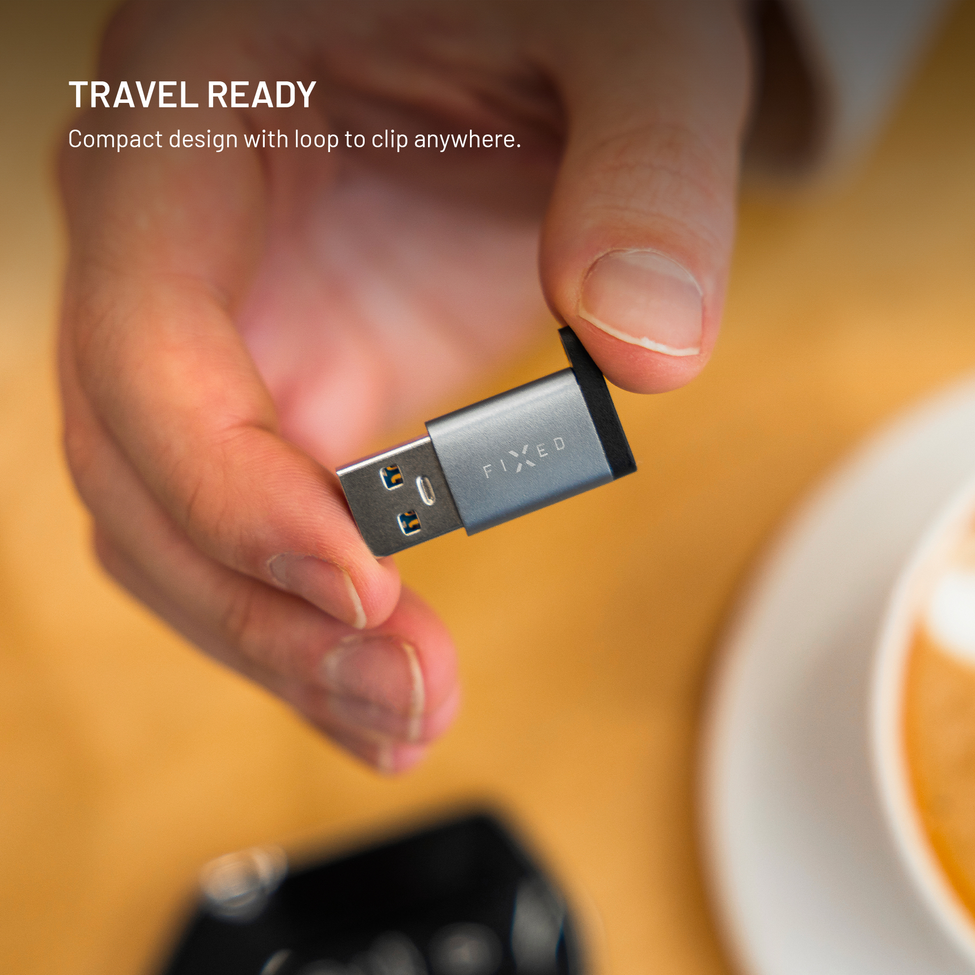Eine Hand hält einen kleinen grauen FIXED USB-Adapter vor einem hölzernen Hintergrund mit einer Tasse.
