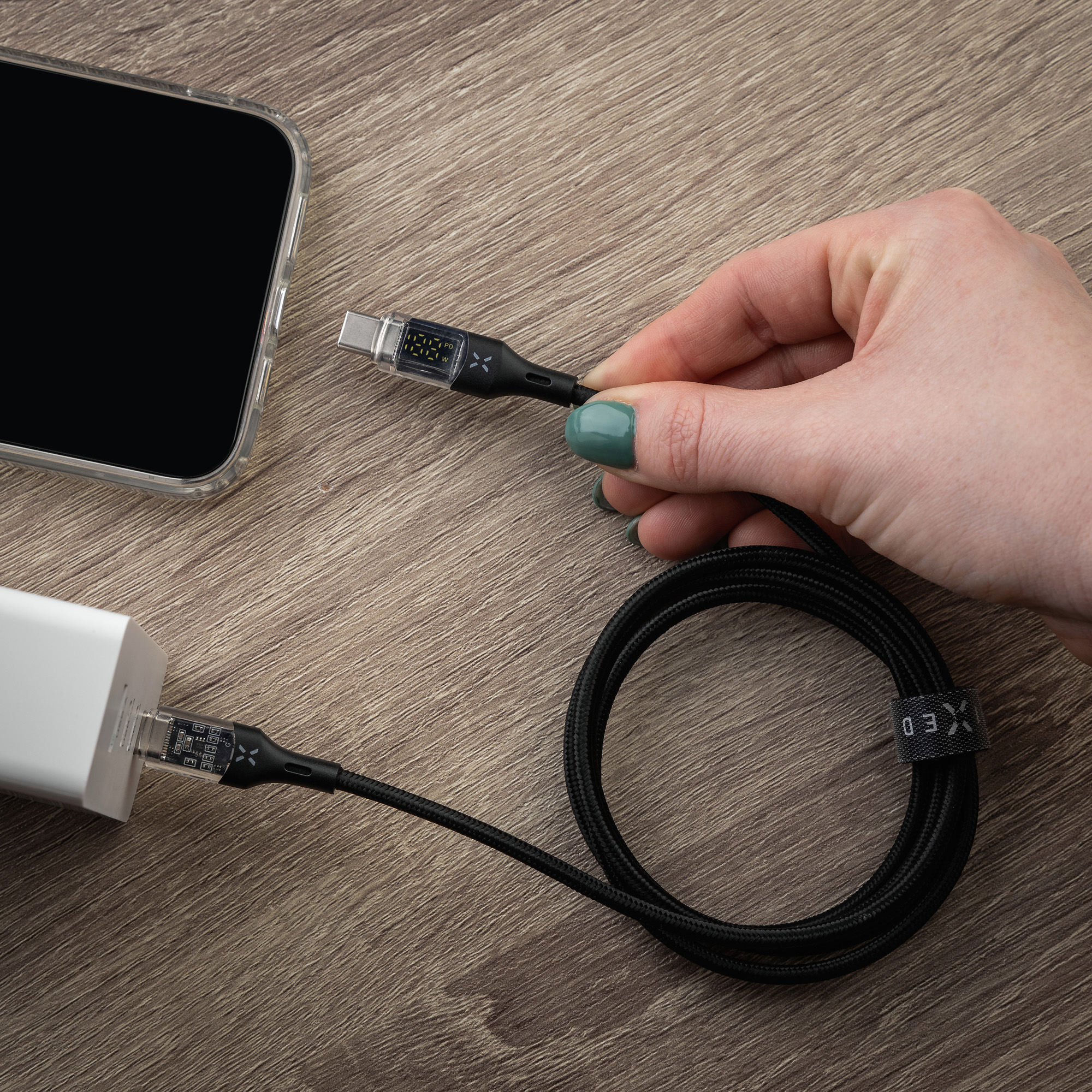 Eine Hand hält ein schwarzes USB-C-Kabel auf Holz, verbunden mit Ladegerät und Handy.