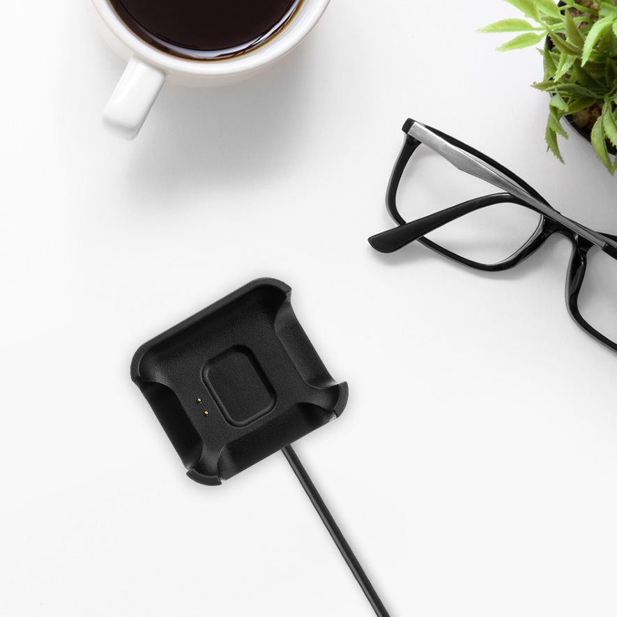 Eine schwarze Smartwatch-Ladestation, eine Brille, eine Kaffeetasse und eine kleine Topfpflanze auf einem weißen Schreibtisch.