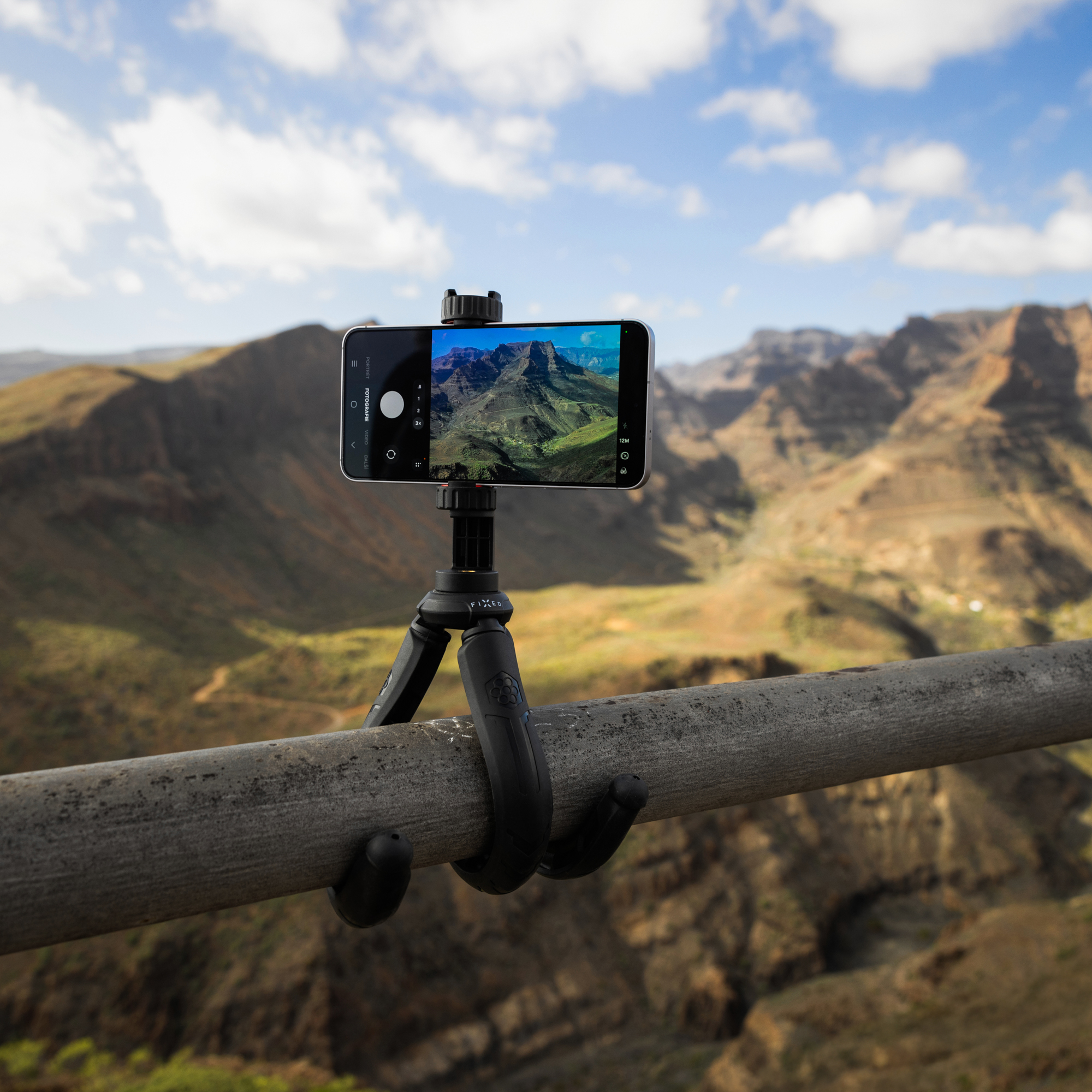 Ein Smartphone auf einem kleinen Stativ, das an einem Metallrohr vor einer bergigen Landschaft befestigt ist.