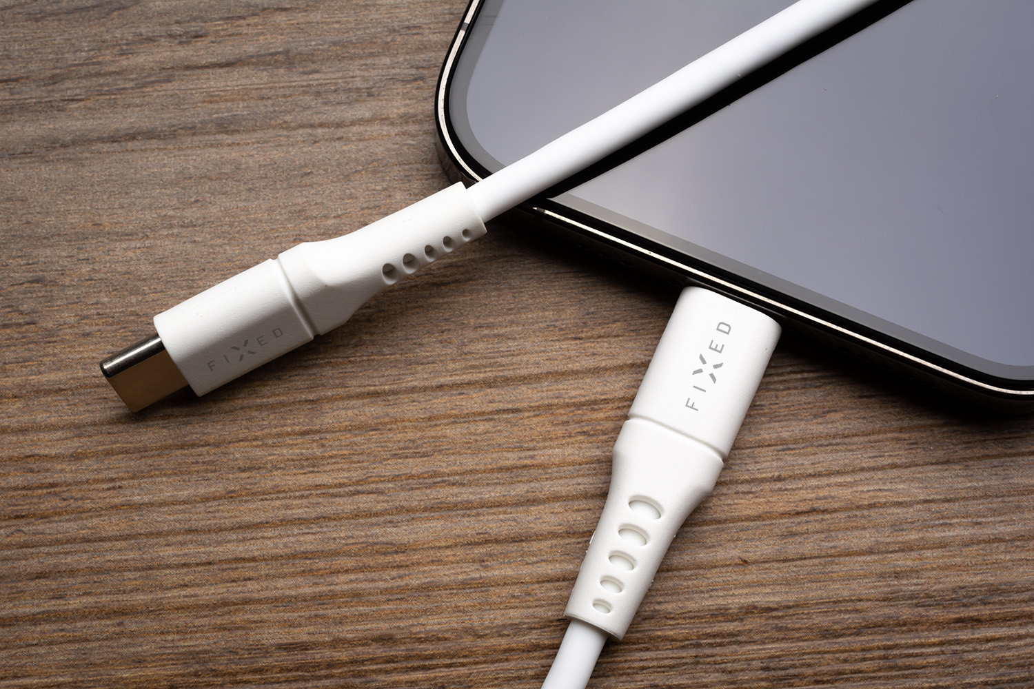 Ein weißes FIXED USB-C-Kabel ist an ein Smartphone angeschlossen, ein zweites liegt daneben auf Holz.