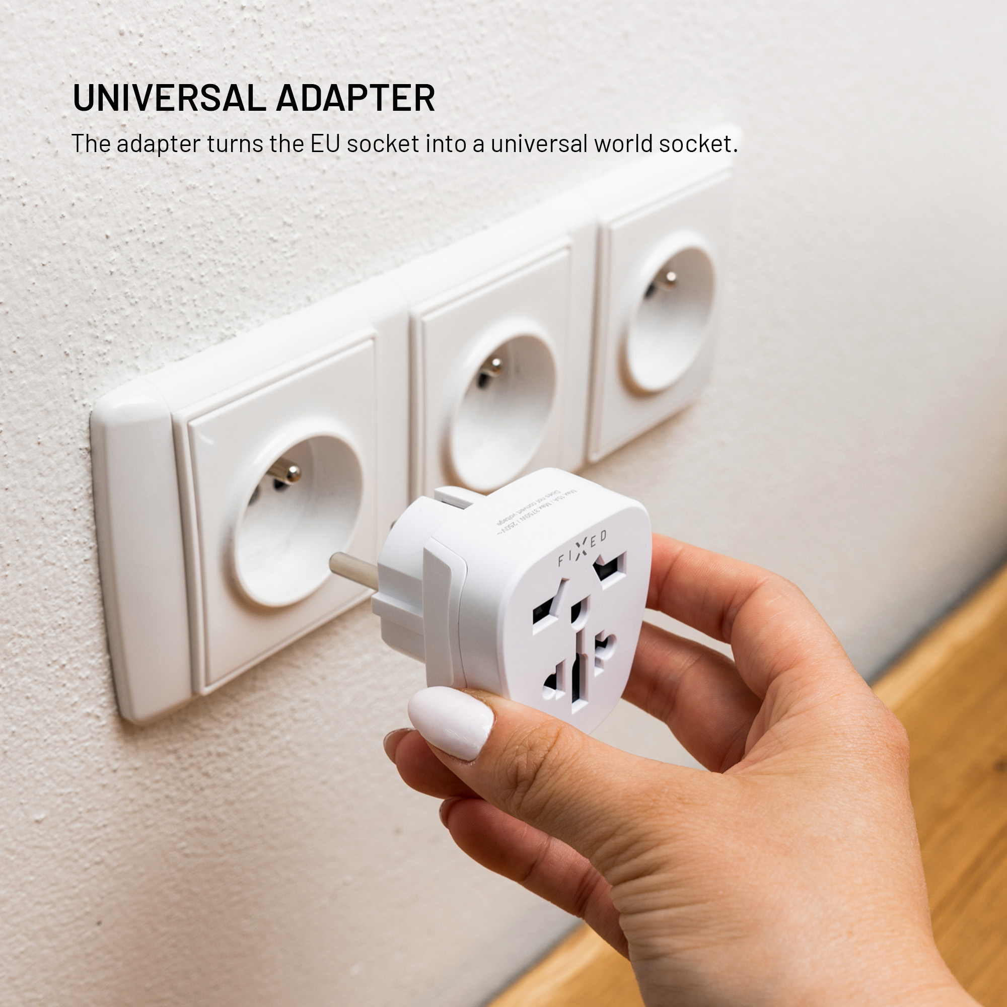 Eine Hand hält einen weißen Universaladapter vor eine Steckdose an einer Wand.