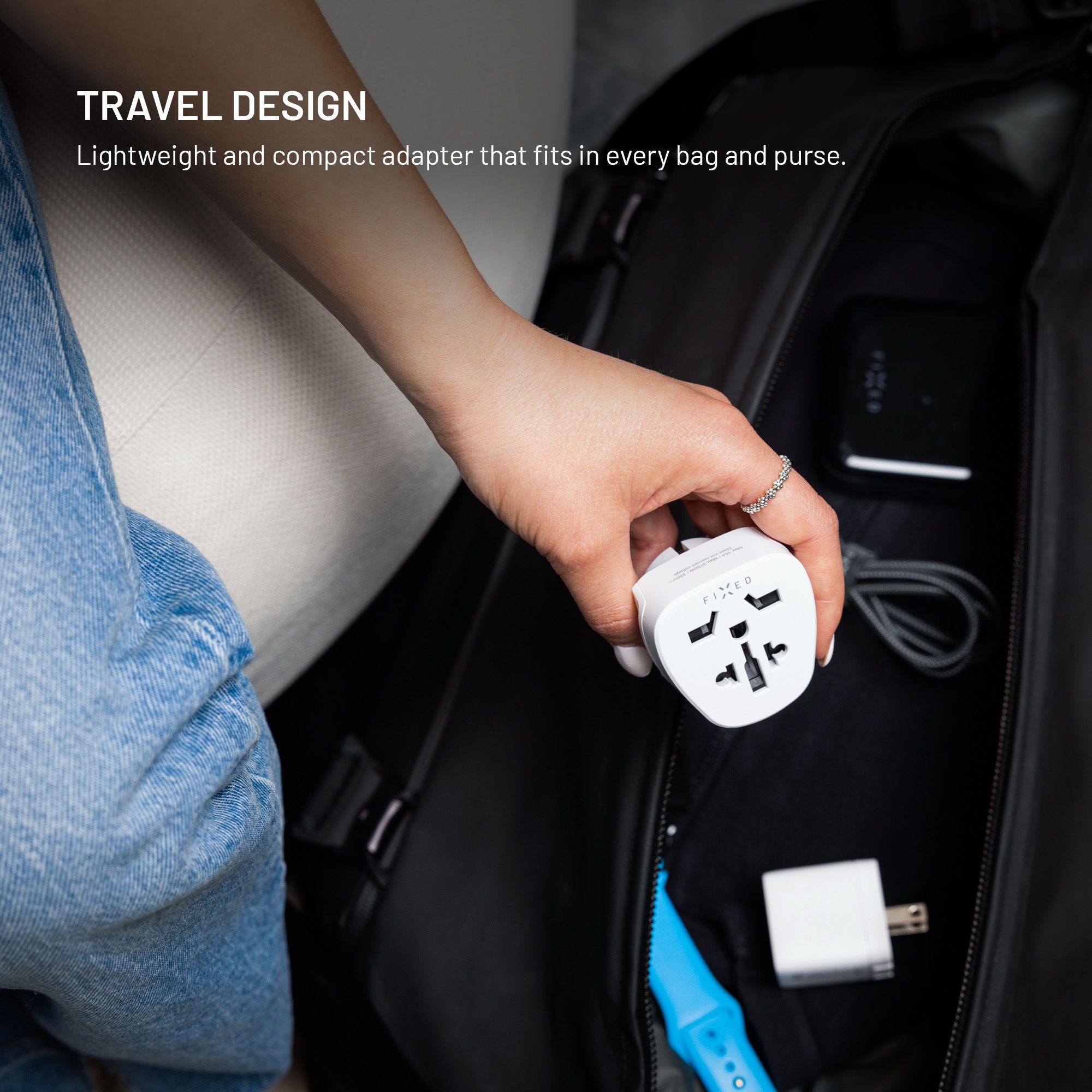 Eine Hand hält einen weißen Reiseadapter über einer schwarzen Tasche. Text lautet: TRAVEL DESIGN.