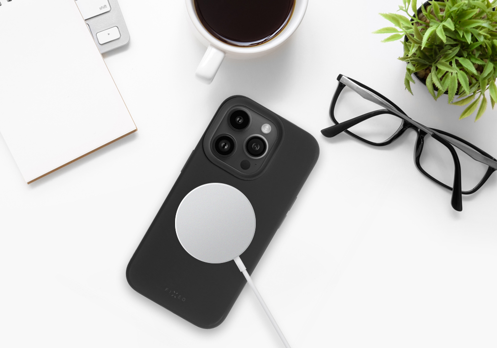 Ein Smartphone mit MagSafe-Ladegerät, Brille, Notizbuch und Kaffeetasse auf weißem Tisch.