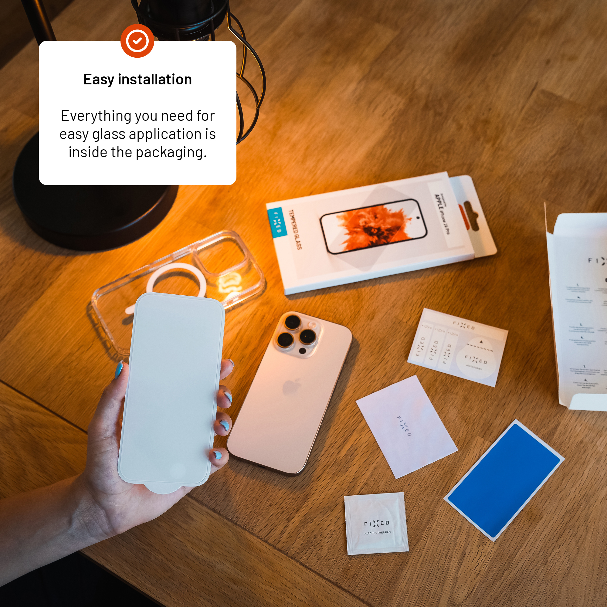 Hand hält Displayschutzglas über einem Holztisch mit iPhone, Hülle und Verpackungszubehör.