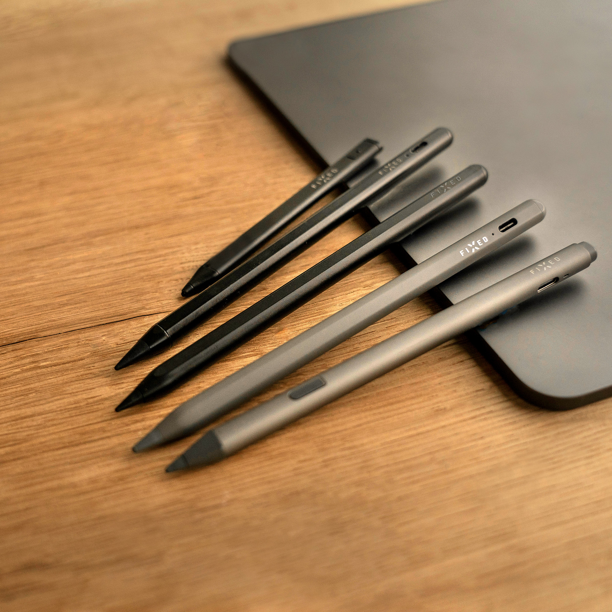 Fünf schwarze Eingabestifte liegen diagonal auf einem Holztisch neben einem dunklen Tablet.