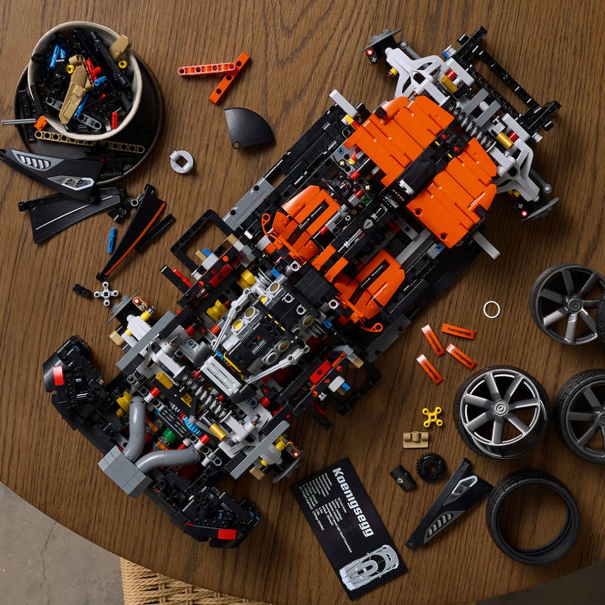 Coche Lego Technic naranja y negro incompleto sobre una mesa de madera, rodeado de piezas y cuatro ruedas.