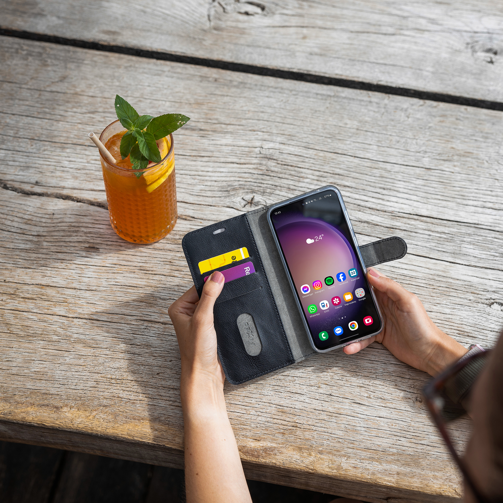 Person hält ein Smartphone im schwarzen Etui auf Holztisch, daneben ein Getränk mit Minze.