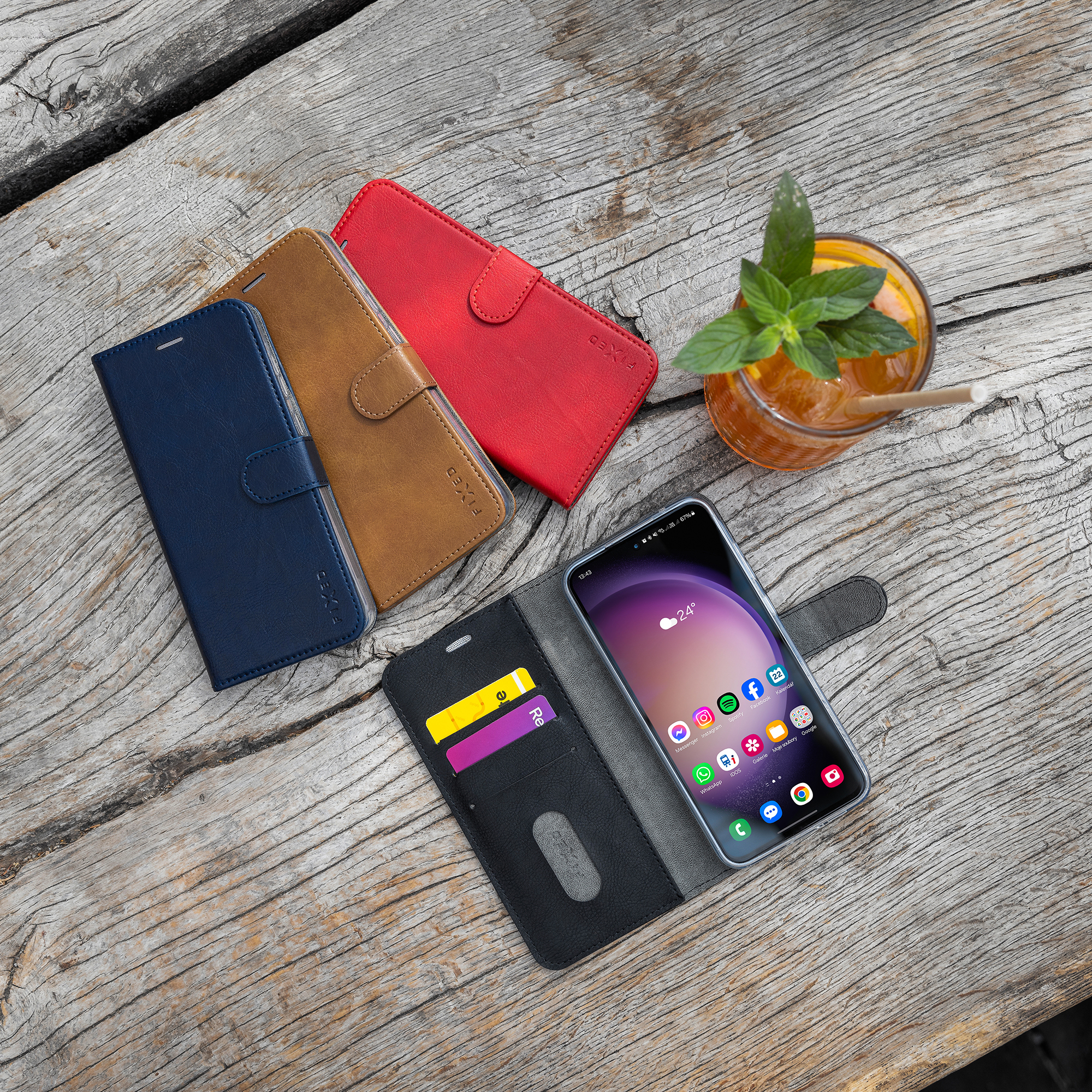 Verschiedenfarbige Leder-Handyhüllen, ein offenes Case mit Smartphone und ein Glas Eistee auf Holz.
