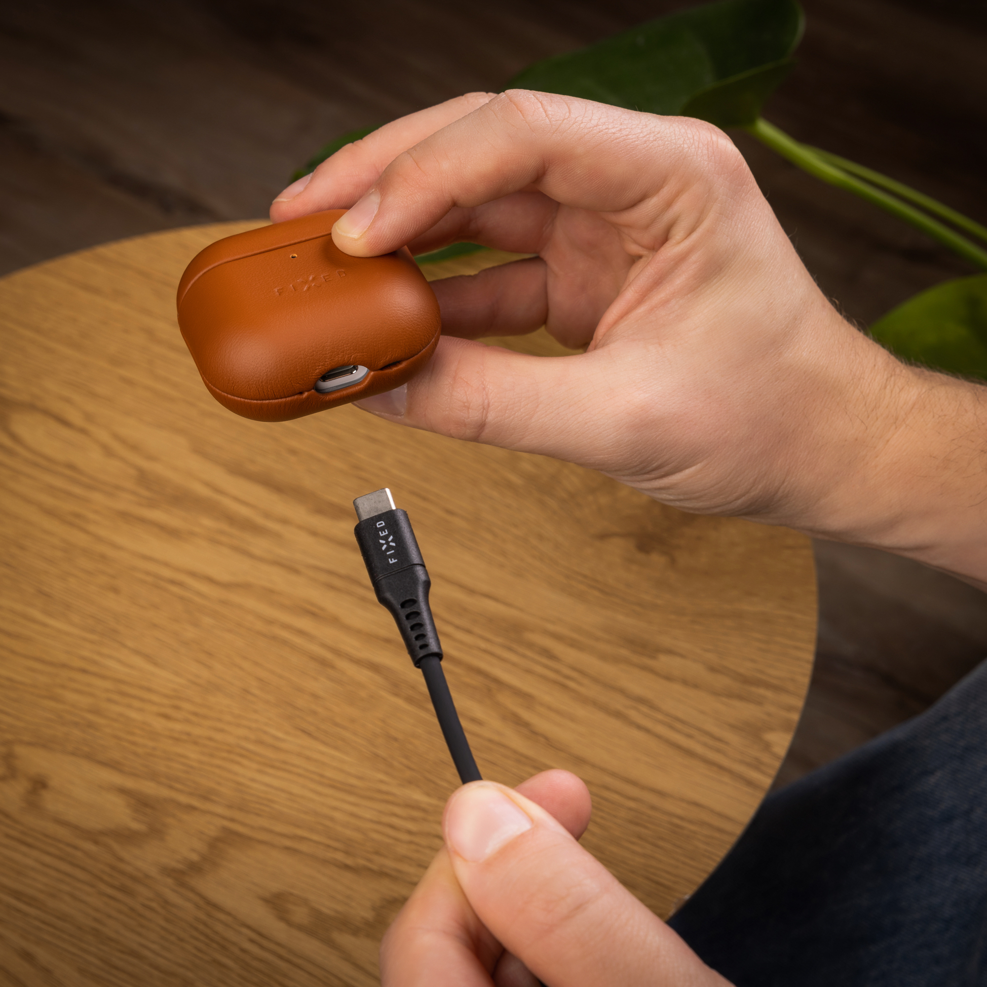 Eine Person hält ein braunes Kopfhörer-Etui über ein schwarzes USB-C-Kabel auf einem Holztisch.