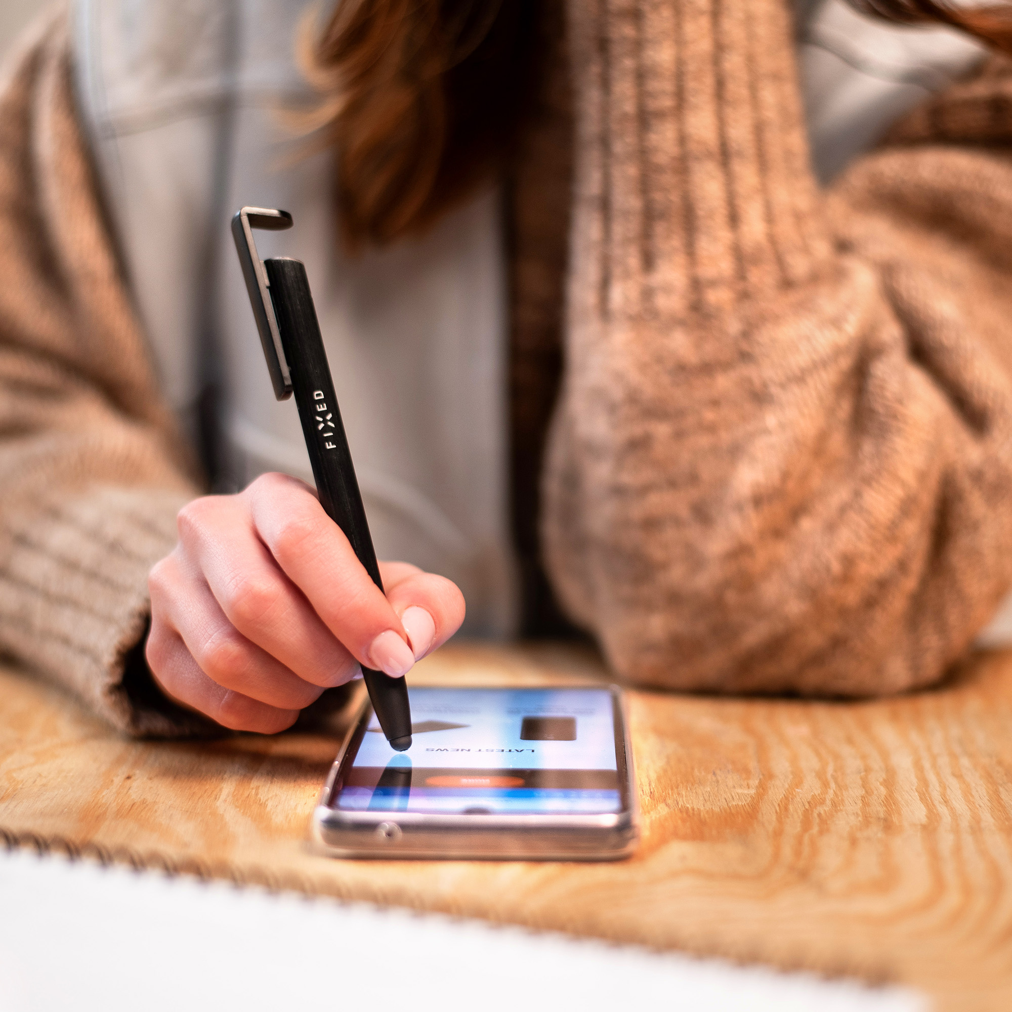 Eine Frau in einem braunen Pullover benutzt einen schwarzen Stift, um auf einem Smartphone zu tippen.