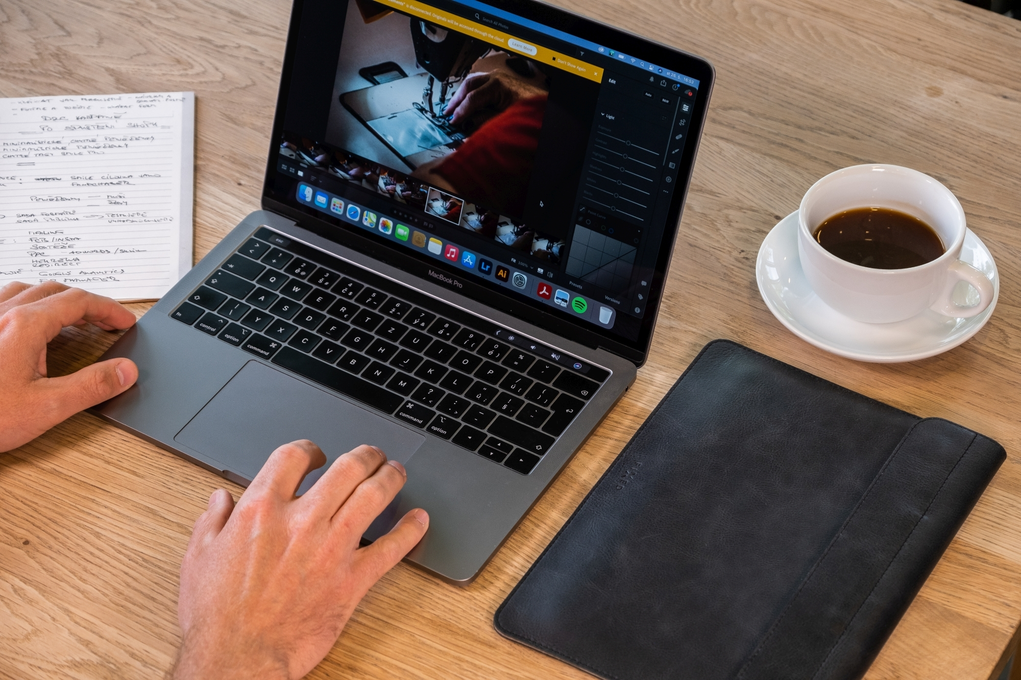 Eine Person benutzt einen Laptop auf einem Holztisch mit Notizbuch, schwarzer Ledertasche und Kaffeetasse.
