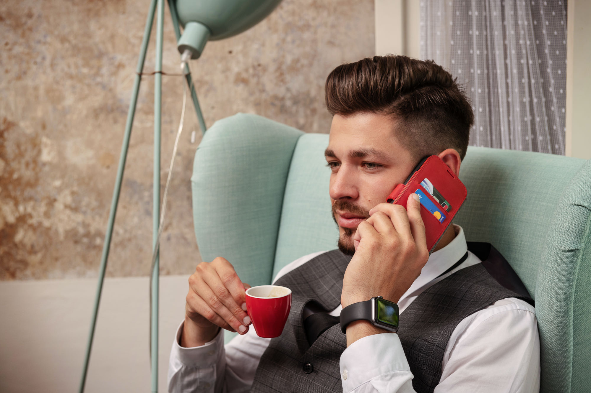 Ein Mann im grauen Anzug sitzt in einem grünen Sessel, telefoniert und hält eine rote Kaffeetasse.