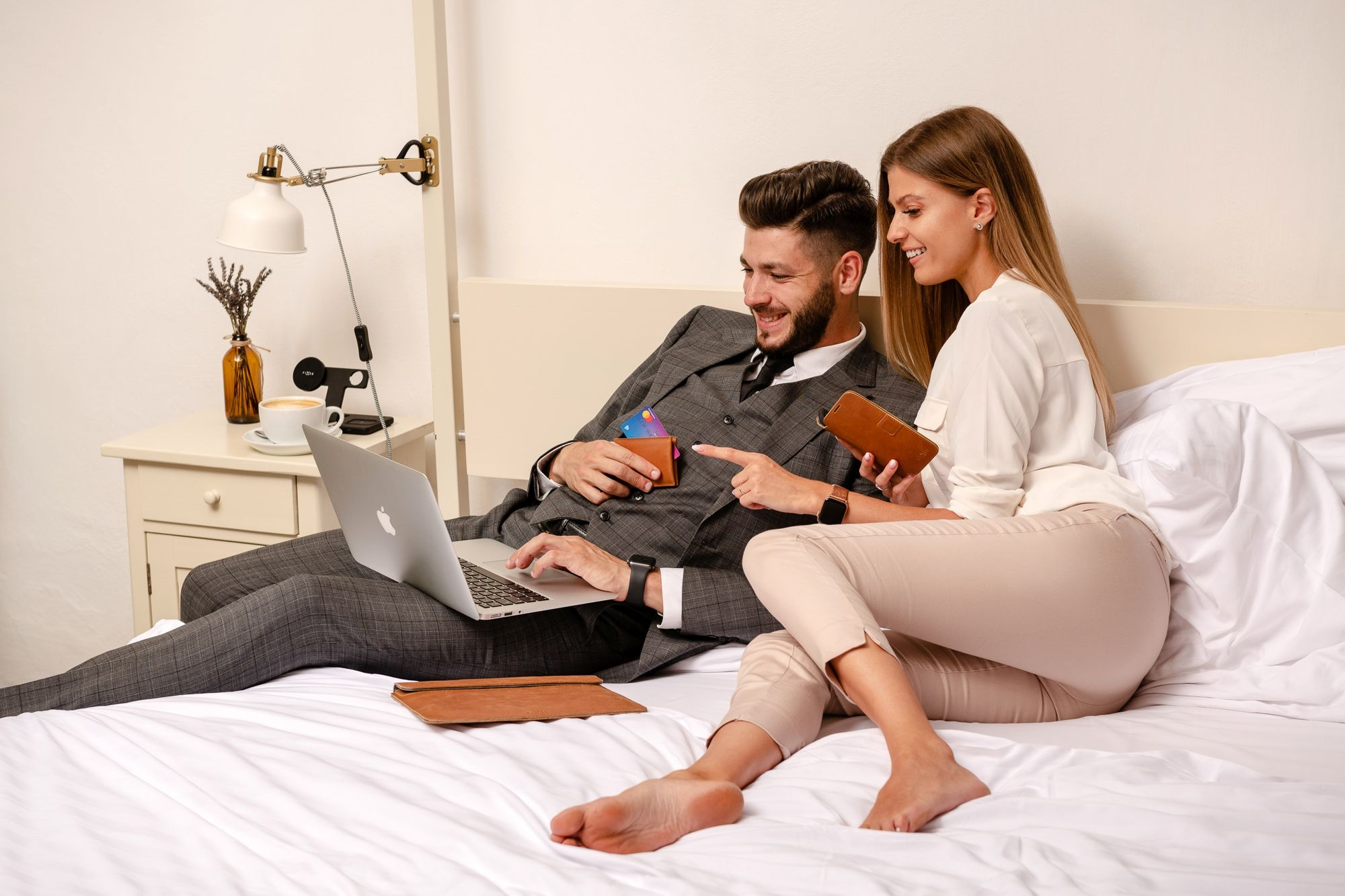 Ein Paar sitzt auf dem Bett; er nutzt einen Laptop, sie hält ein Portemonnaie und zeigt darauf.