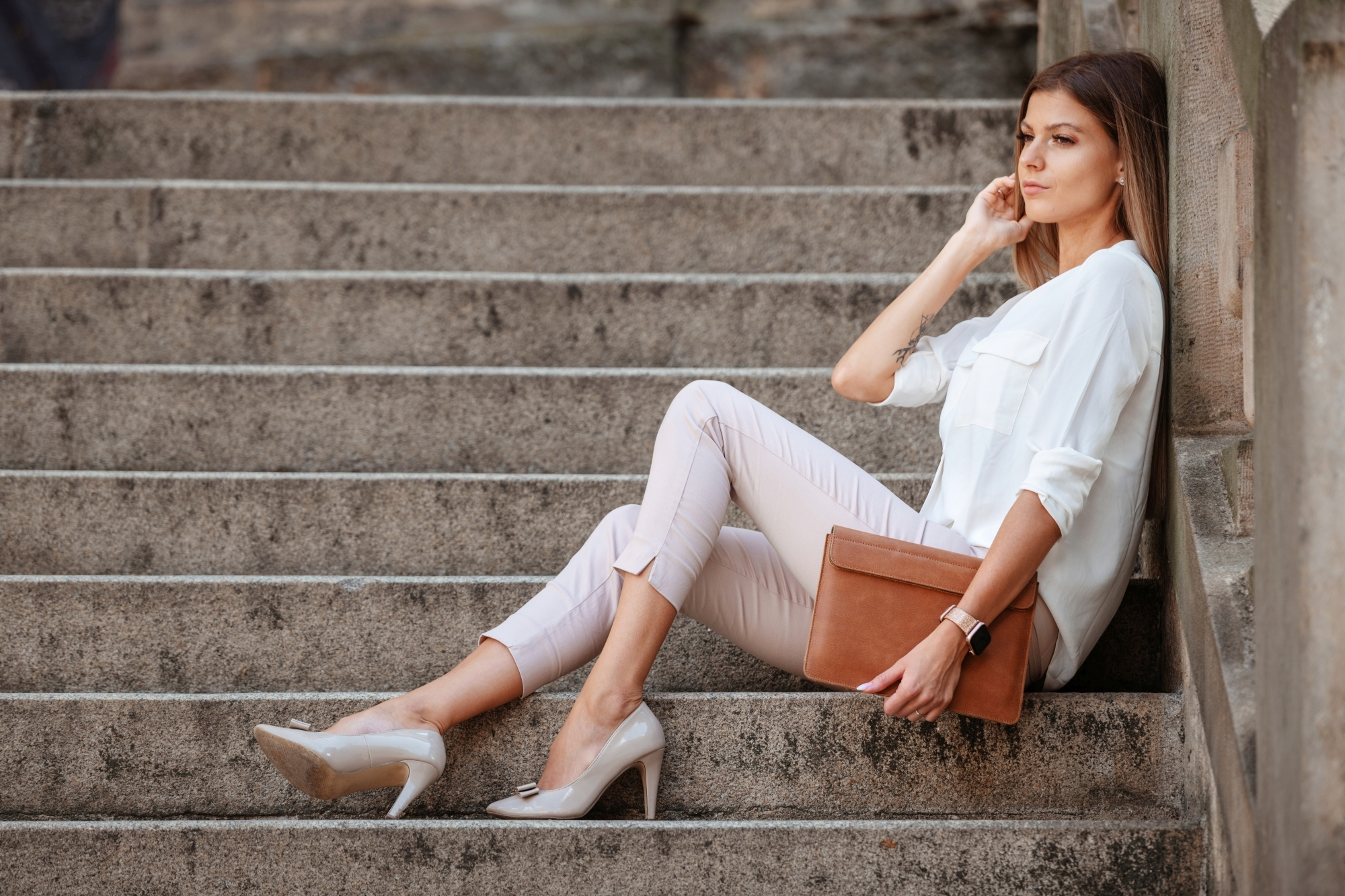 Eine Frau in weißer Bluse und hellen Hosen sitzt auf Steintreppen und hält eine braune Tasche.