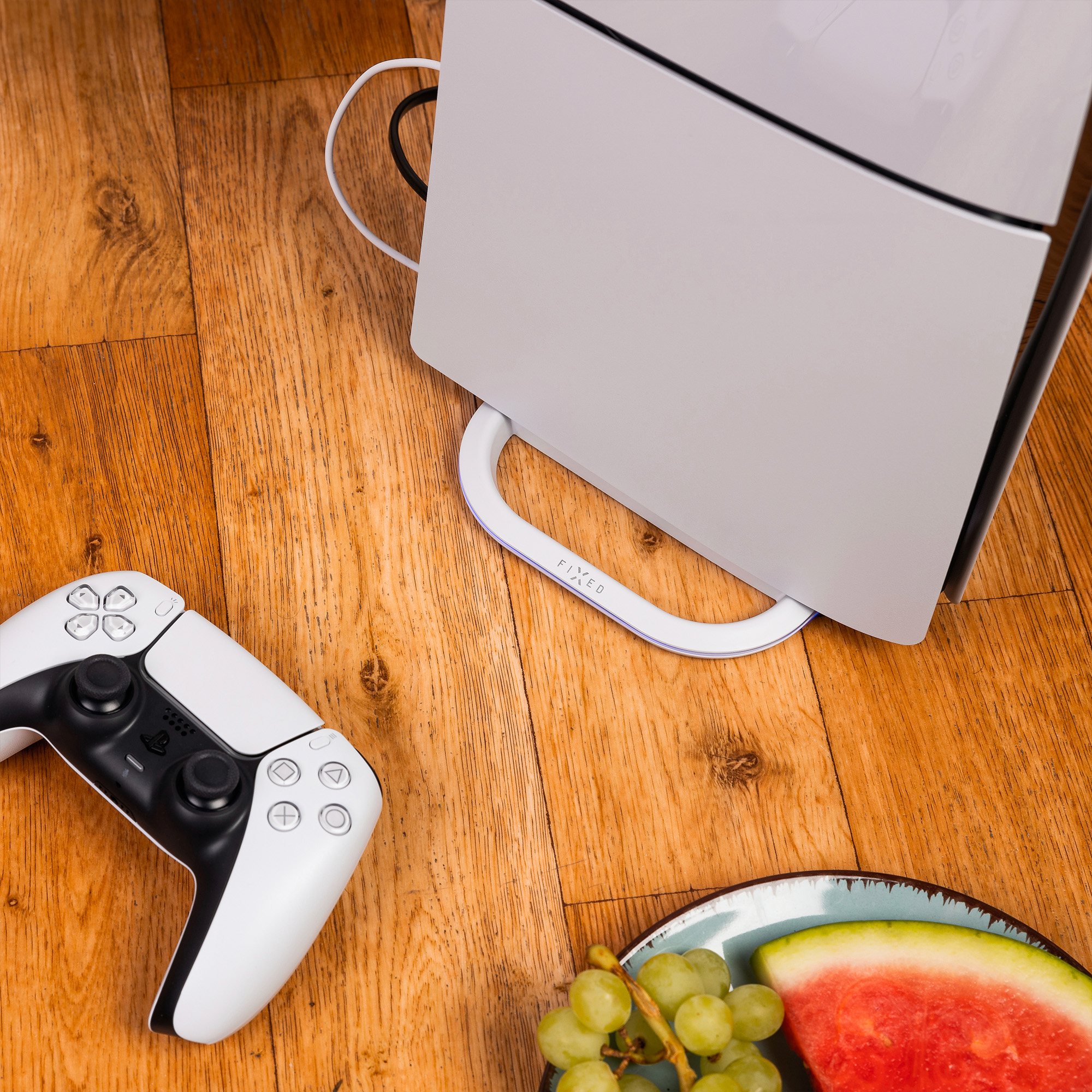 Eine weiße Spielkonsole, ein Controller und ein Teller mit Trauben und Wassermelone auf Holzboden.