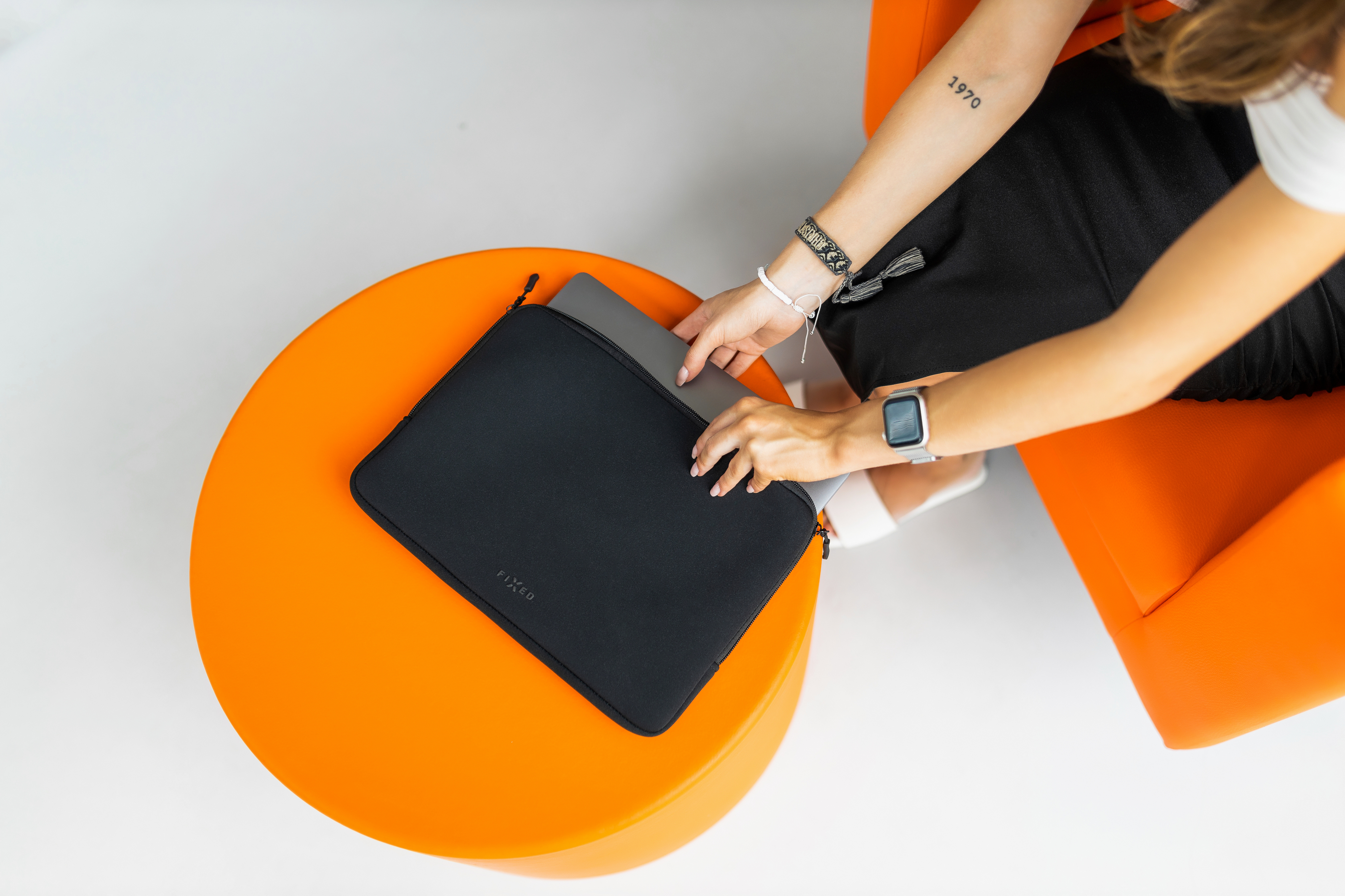 Eine Frau in einem orangefarbenen Sessel legt einen Laptop in eine schwarze Schutzhülle auf einem orangefarbenen Tisch.