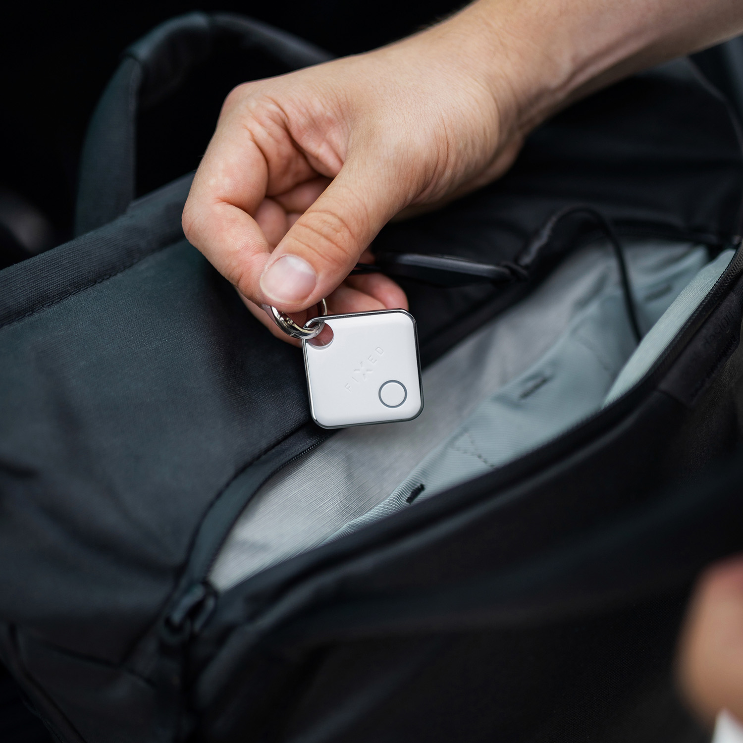 Eine Hand hält einen kleinen, weißen Smart-Tracker an einem Schlüsselring über die offene Tasche eines schwarzen Rucksacks.