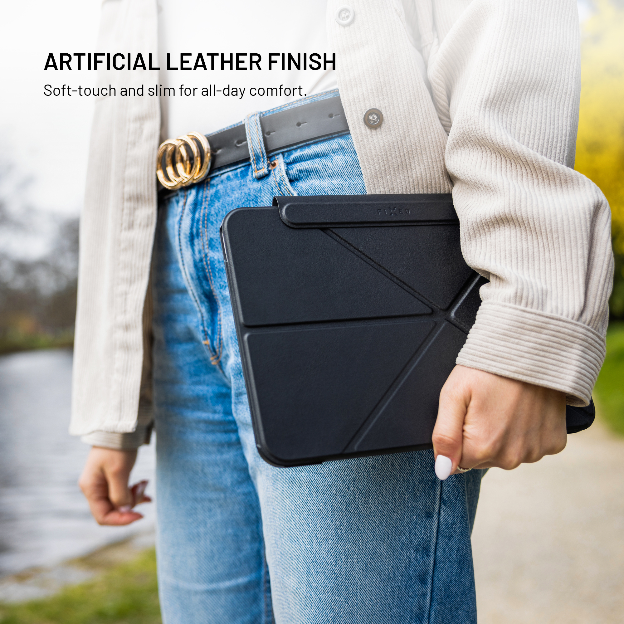 Person in Jeans und hellem Mantel hält ein schwarzes Tablet mit Ledercase outdoors.