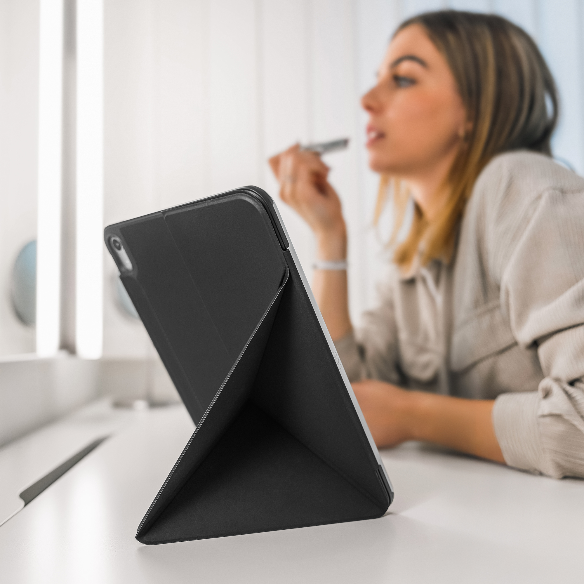 Ein schwarzes Tablet im Ständer steht auf dem Tisch, während eine Frau im Hintergrund Lippenstift aufträgt.