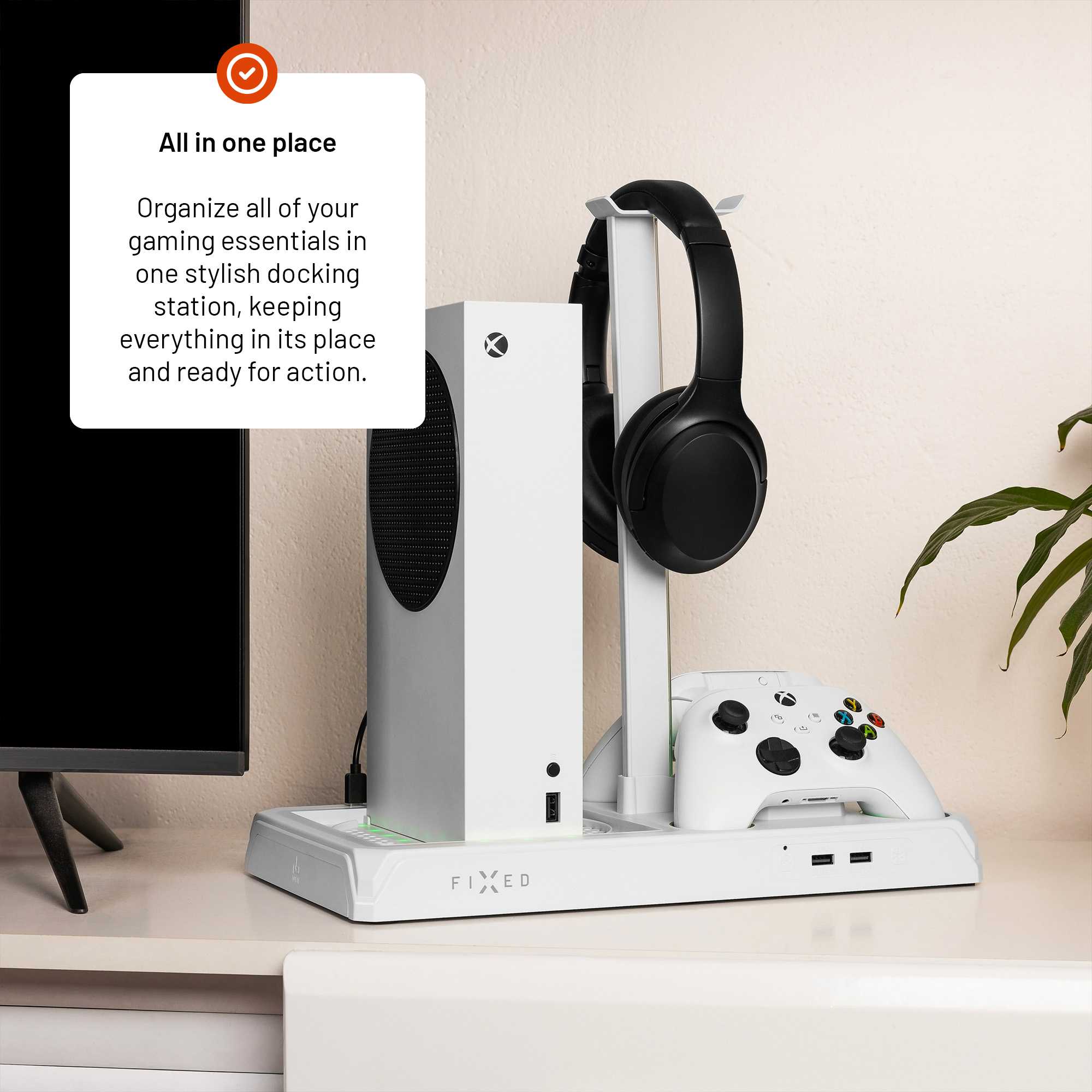 Eine weiße Gaming-Dockingstation hält eine Xbox, einen Controller und schwarze Kopfhörer neben einem Fernseher.