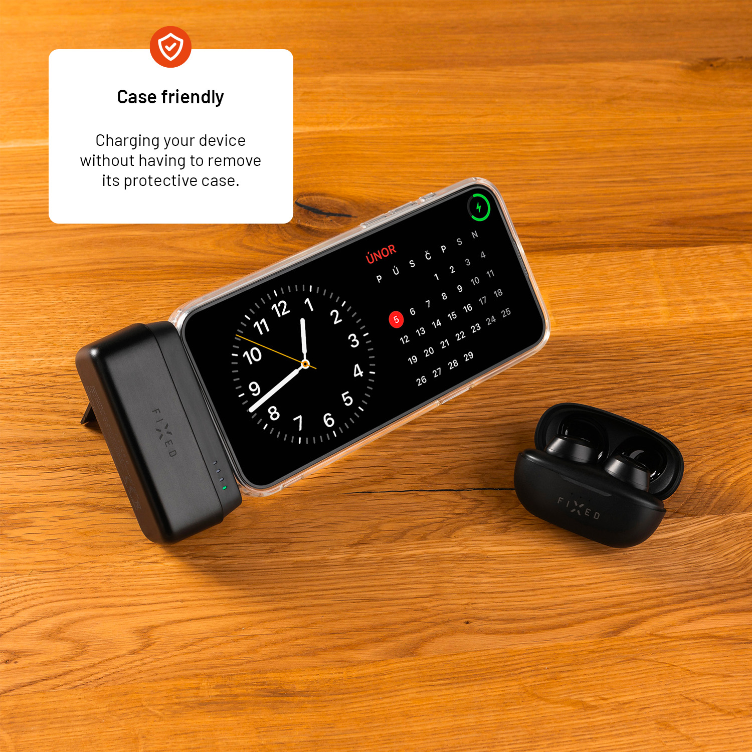 Uno smartphone in custodia si ricarica su un supporto, con auricolari neri accanto su legno.