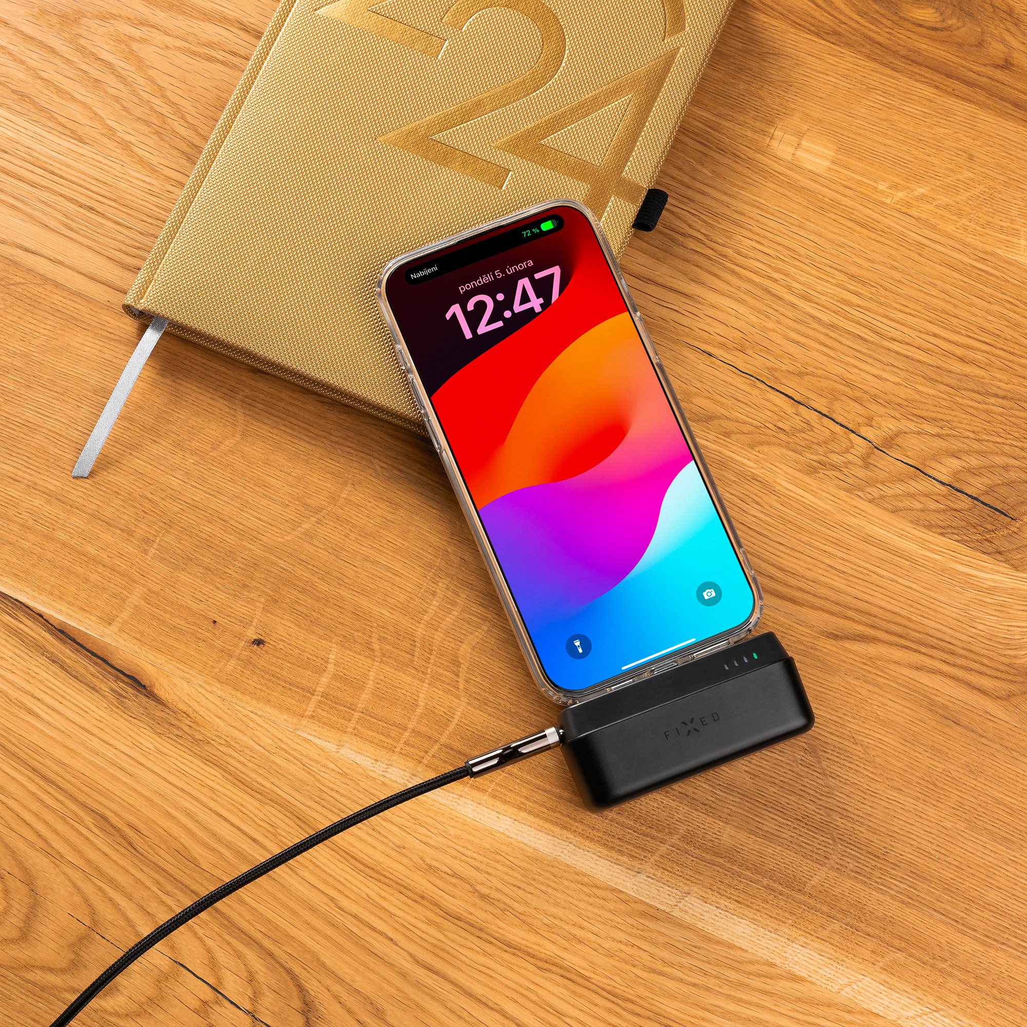 Uno smartphone si ricarica su un power bank su un tavolo di legno accanto a un taccuino dorato.