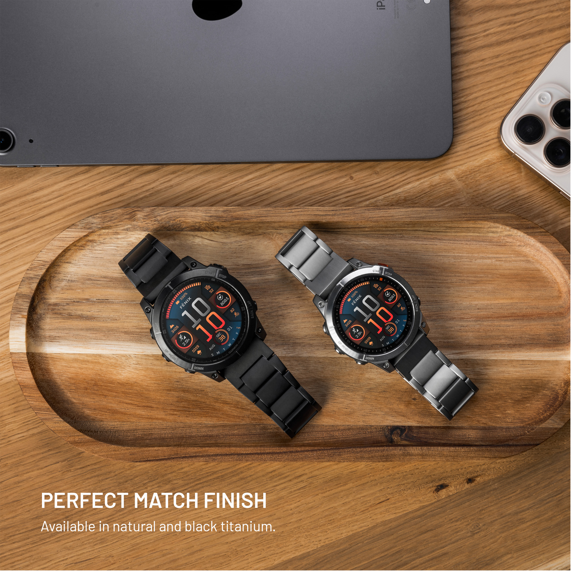 Zwei Smartwatches aus Titan in Schwarz und Silber liegen auf einem Holztablett, umgeben von einem Tablet und einem Smartphone.