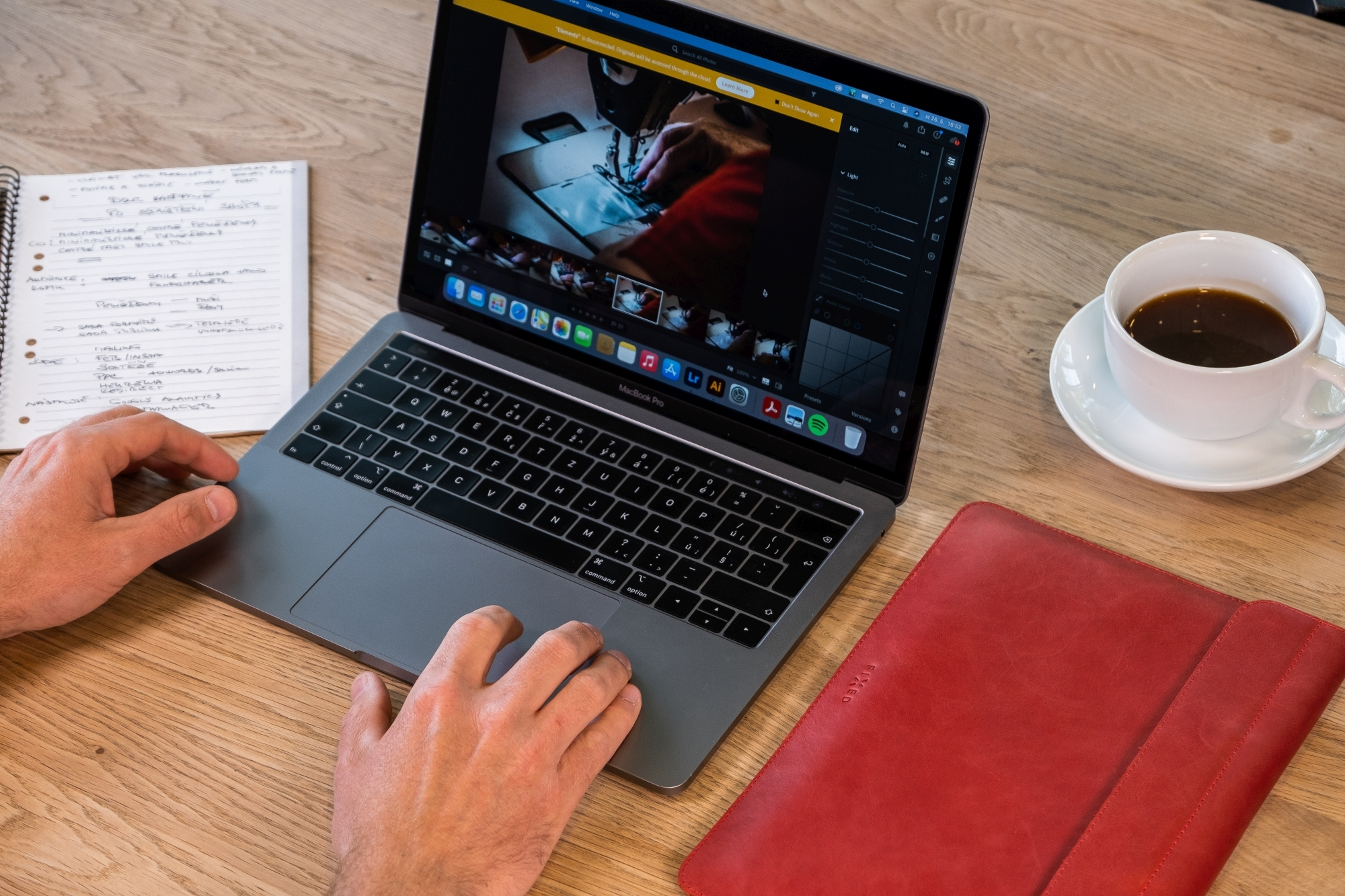 Eine Person nutzt einen Laptop auf einem Holztisch mit Notizbuch, Kaffeetasse und roter Ledertasche.