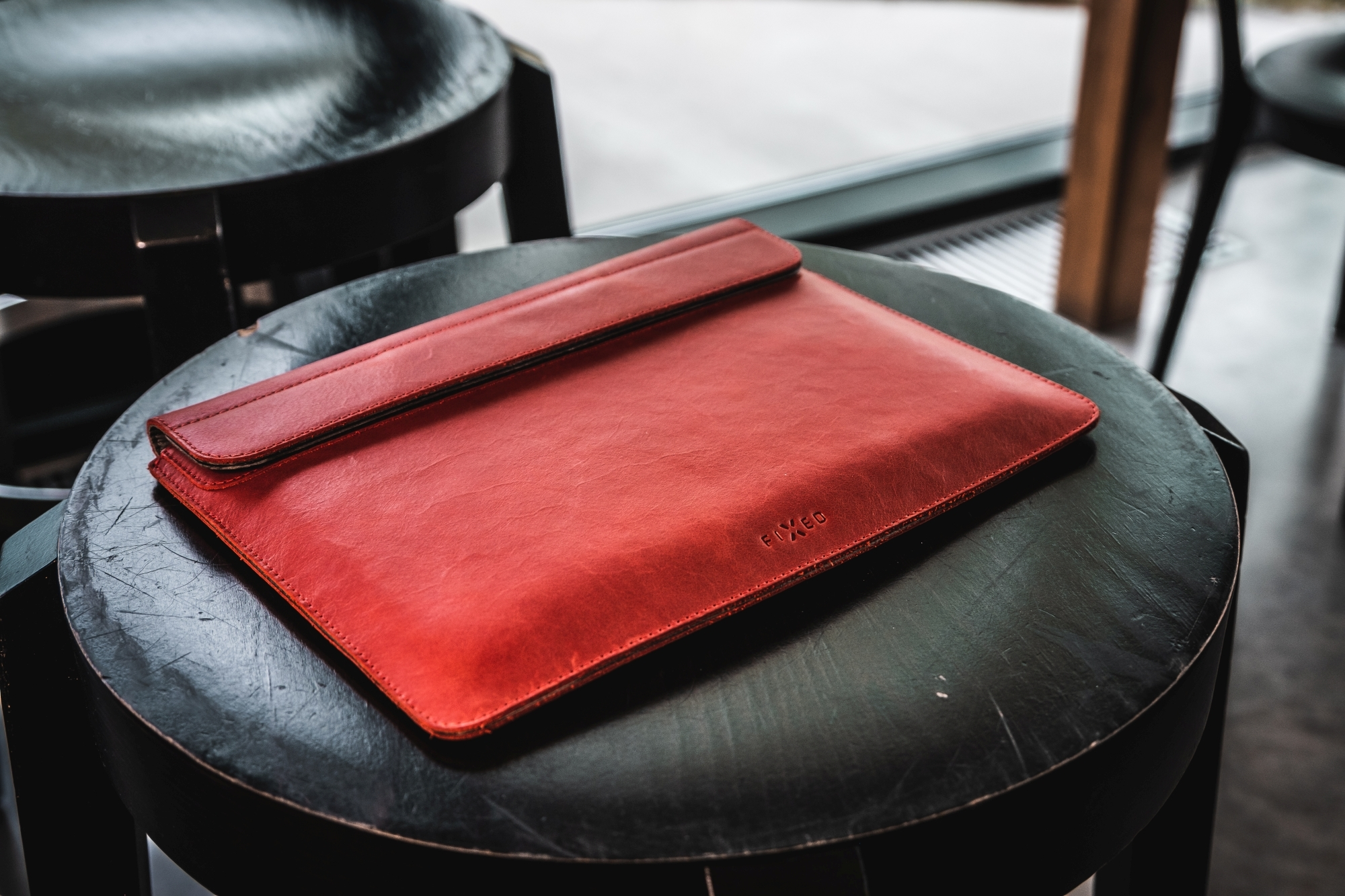 Eine rote Laptophülle aus Leder liegt drinnen auf einem runden Hocker, mit Möbeln und einem Fenster im Hintergrund.