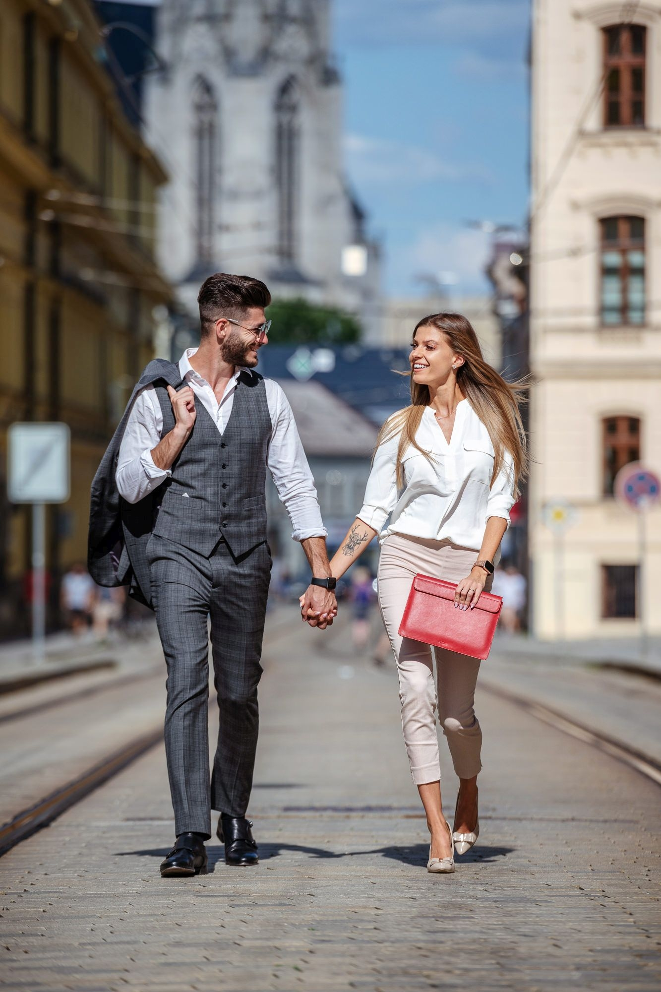 Ein Paar hält Händchen und geht eine städtische Straße entlang, er trägt einen Anzug und sie hält eine rote Tasche.