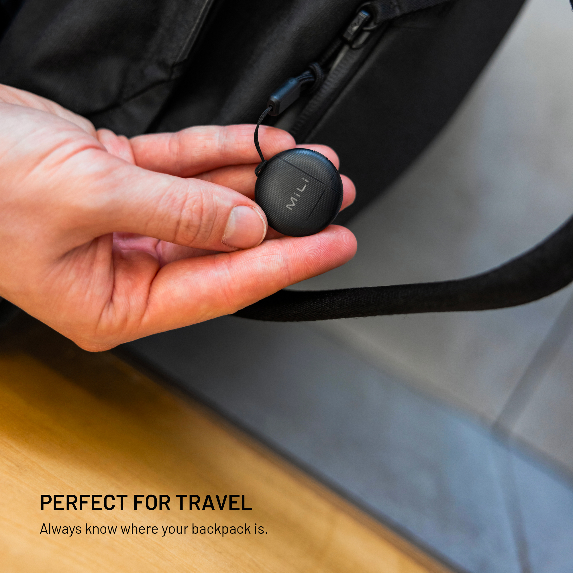 Hand hält einen kleinen schwarzen MiLi-Tracker, befestigt am Reißverschluss eines Rucksacks.