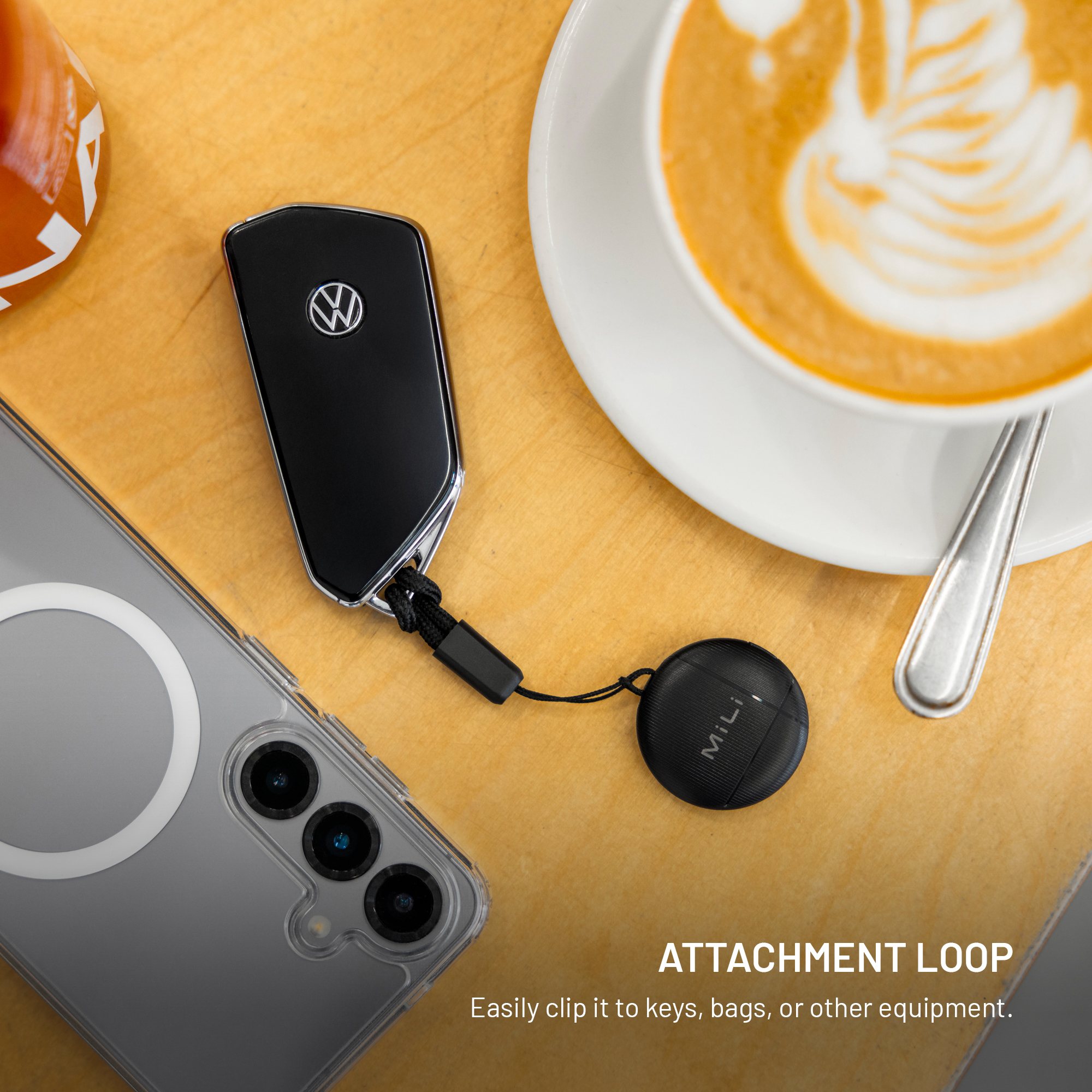 Ein Volkswagen-Autoschlüssel mit einem runden MiLi-Anhänger liegt auf einem Holztisch neben einer Tasse Cappuccino und einem Smartphone.