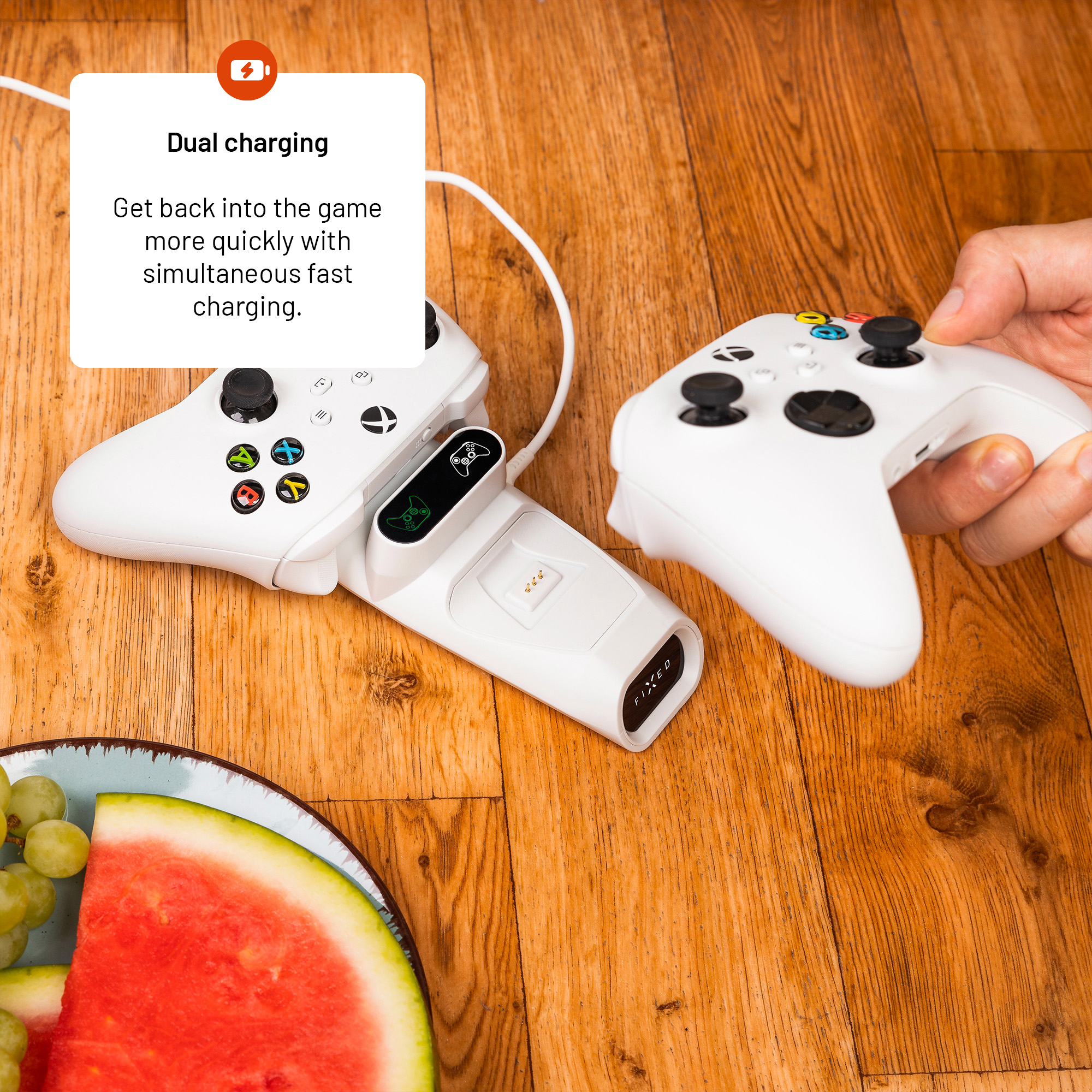 Eine Hand hält einen weißen Xbox-Controller an einer Ladestation auf Holztisch mit Wassermelone.