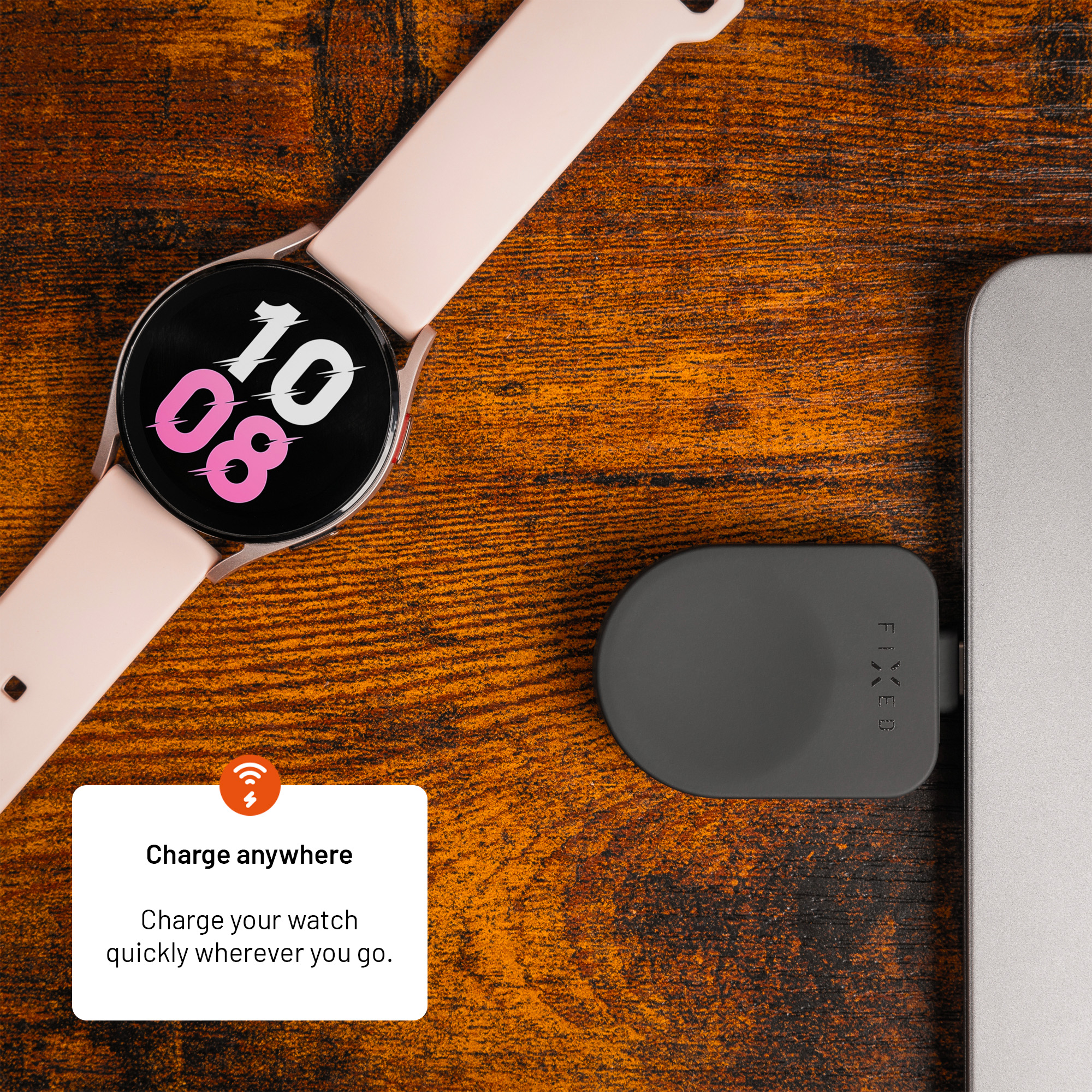 Eine rosa Smartwatch mit Digitaldisplay liegt auf einer Holzoberfläche neben einem schwarzen Ladegerät an einem Laptop.