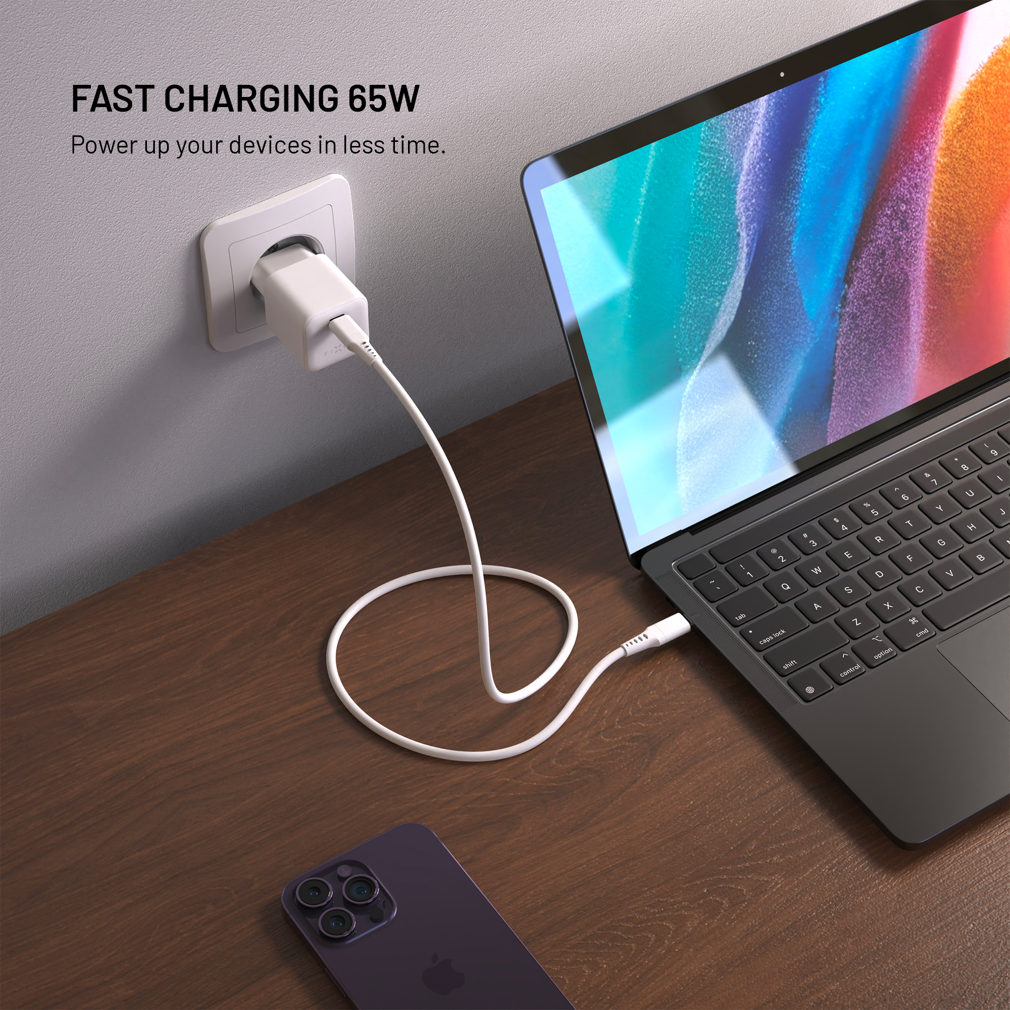 Ein 65W-Ladegerät in einer Steckdose lädt einen Laptop auf einem Holztisch mit einem Smartphone daneben.