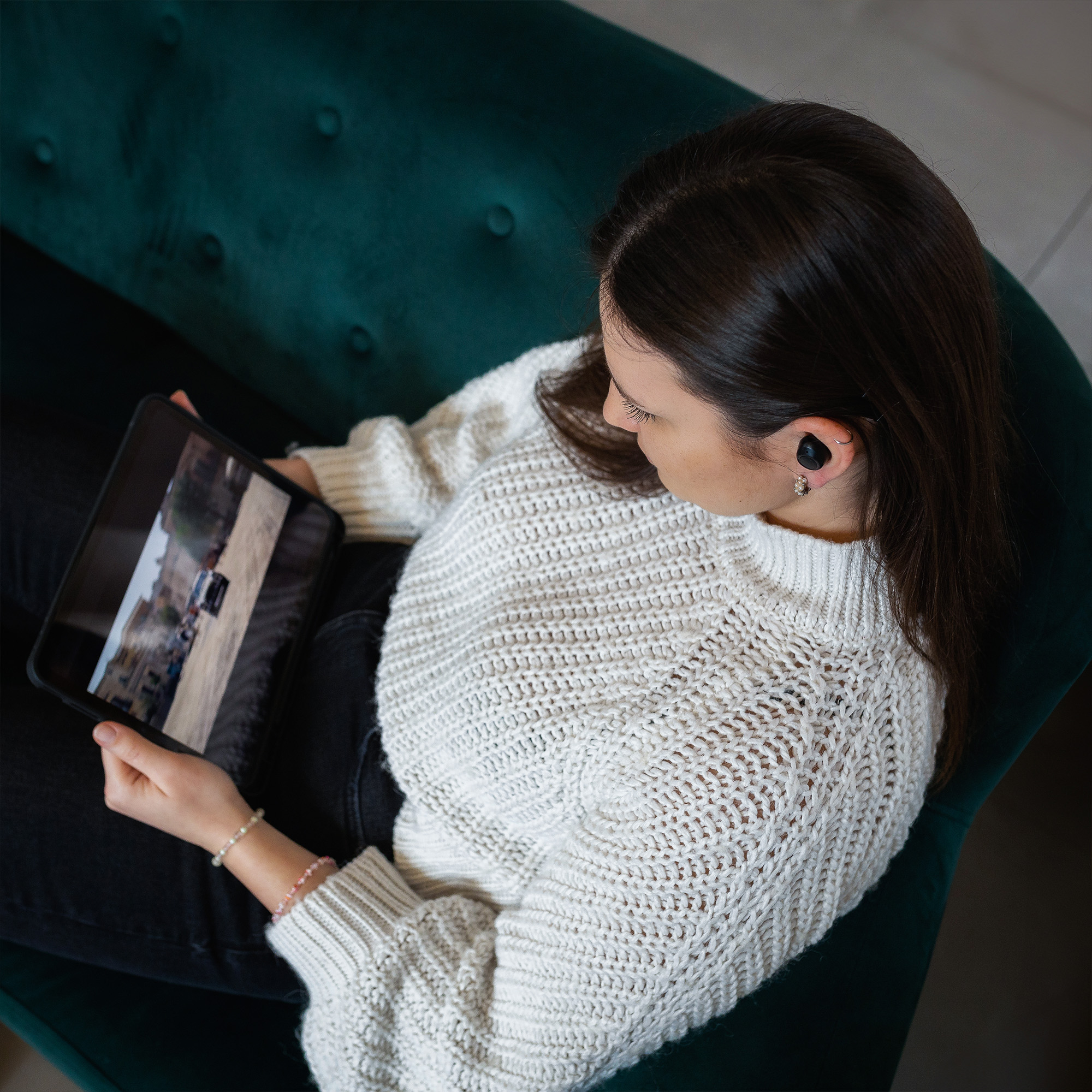 Eine Frau im weißen Pullover sitzt auf einem grünen Sofa und hält ein Tablet mit einem Video.