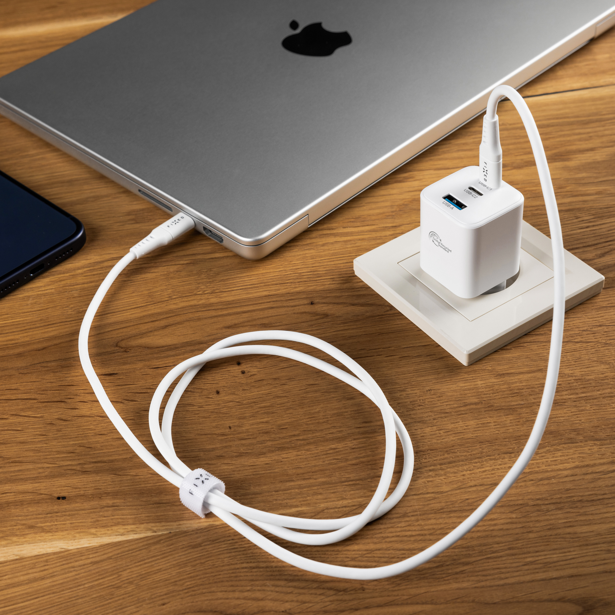 Ein weißes Ladegerät in einer Steckdose ist über ein Kabel mit einem grauen Laptop auf einem Holztisch verbunden.