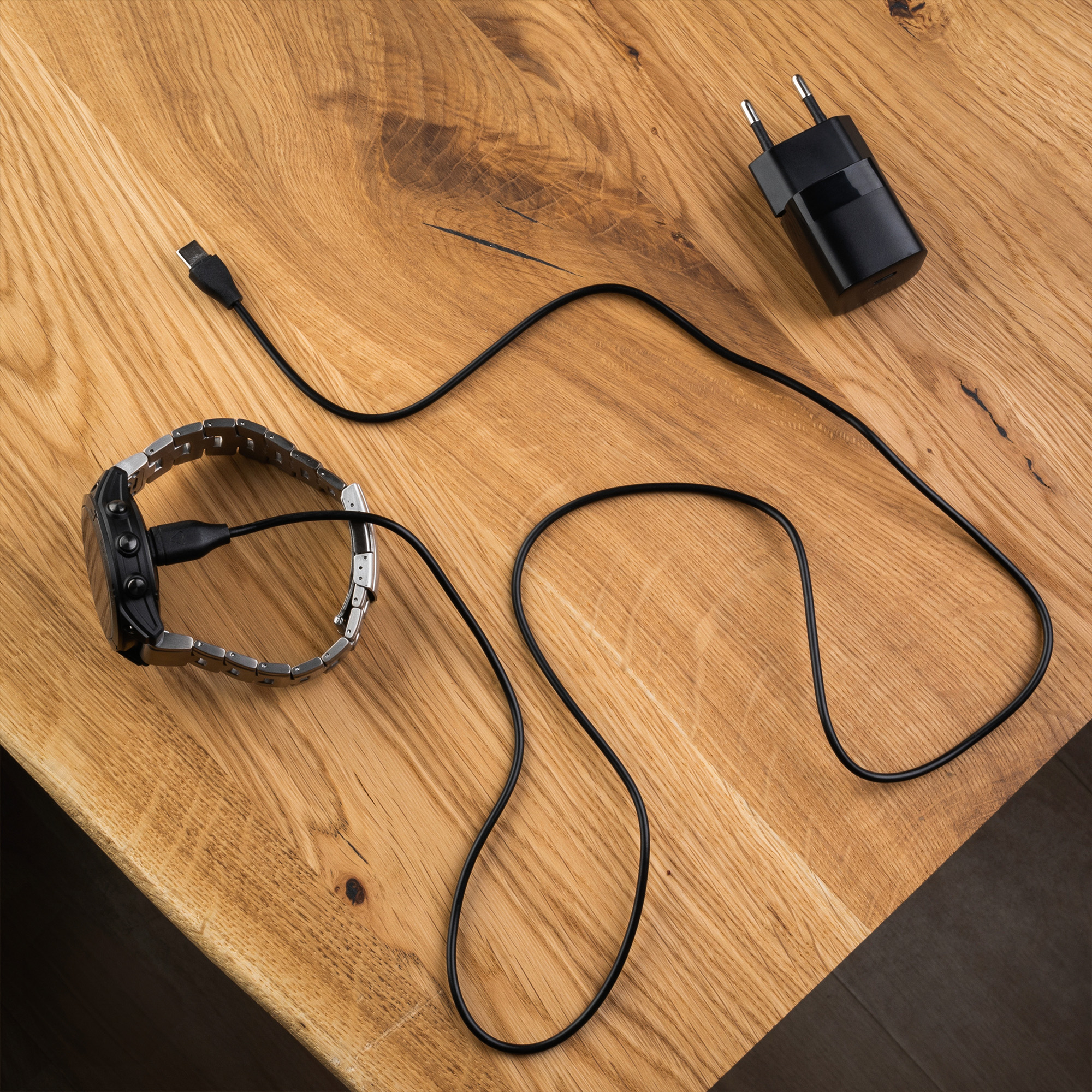 Eine Smartwatch, die mit einem schwarzen USB-Kabel und einem Wandladegerät verbunden ist, auf einem Holztisch.