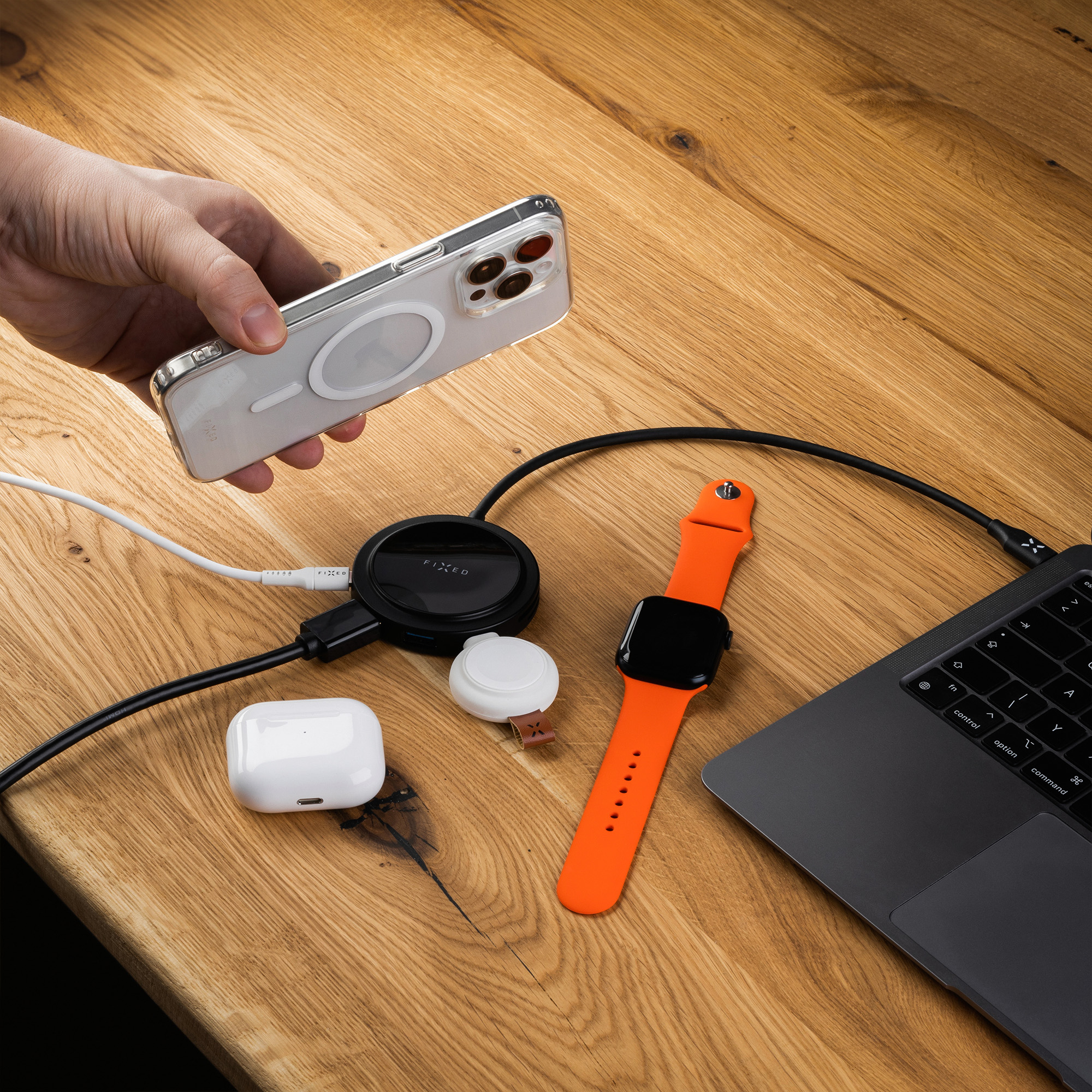 Hand hält eine transparente Handyhülle über eine Ladestation auf Holz, daneben Apple Watch und Laptop.