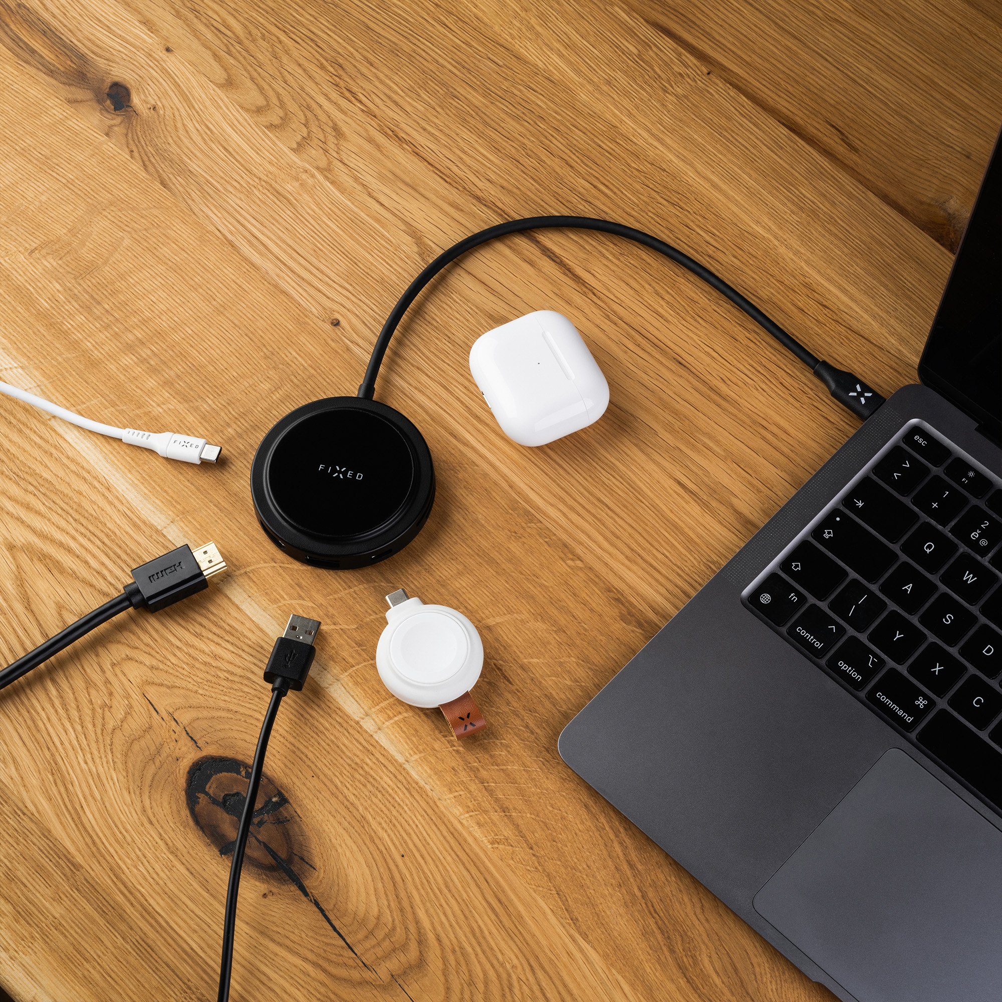Ein Laptop, ein runder schwarzer Hub, AirPods, Kabel und ein Ladegerät auf einer Holzoberfläche.