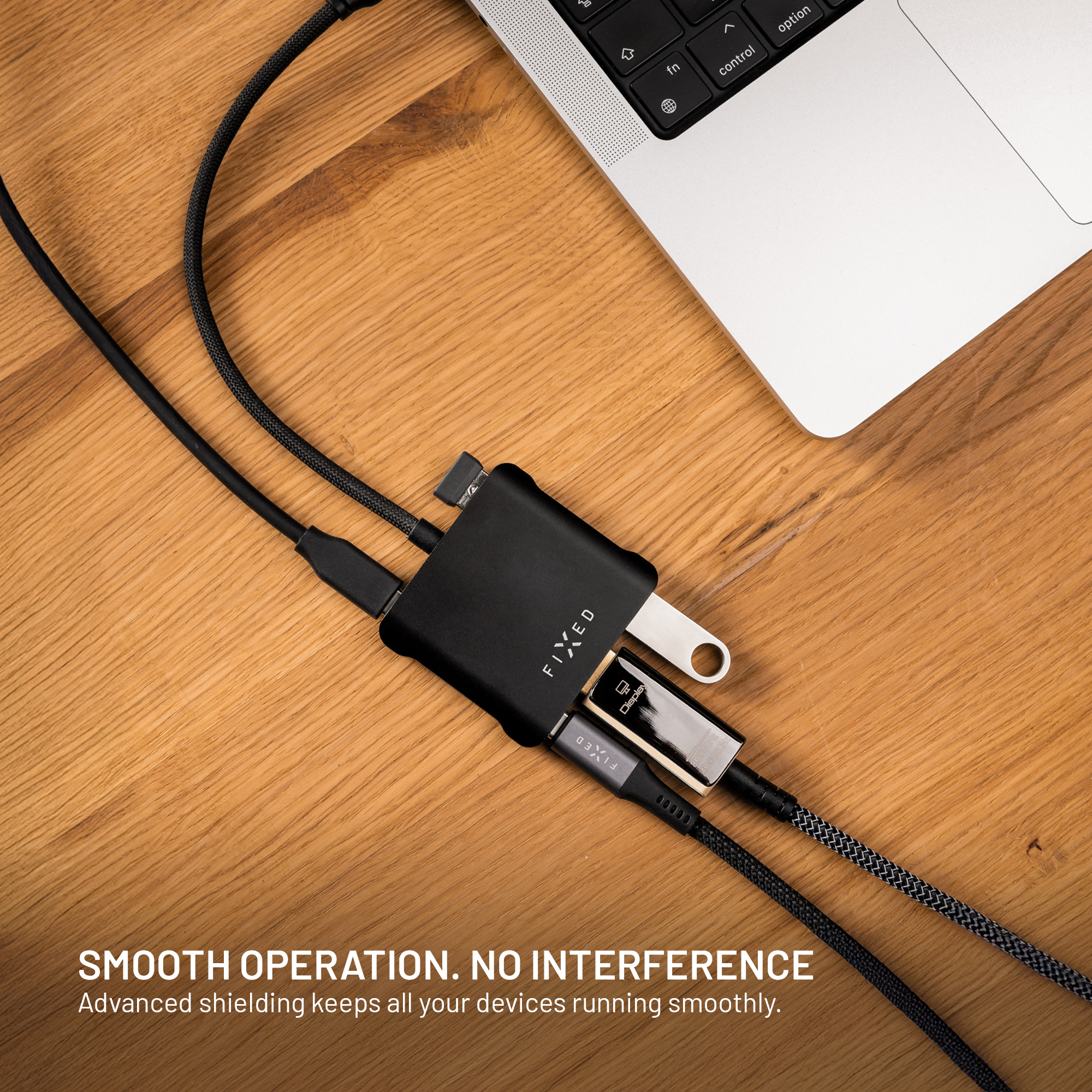 Ein schwarzer USB-C-Hub mit angeschlossenen Kabeln und Adaptern liegt auf einem Holztisch neben einem Laptop.