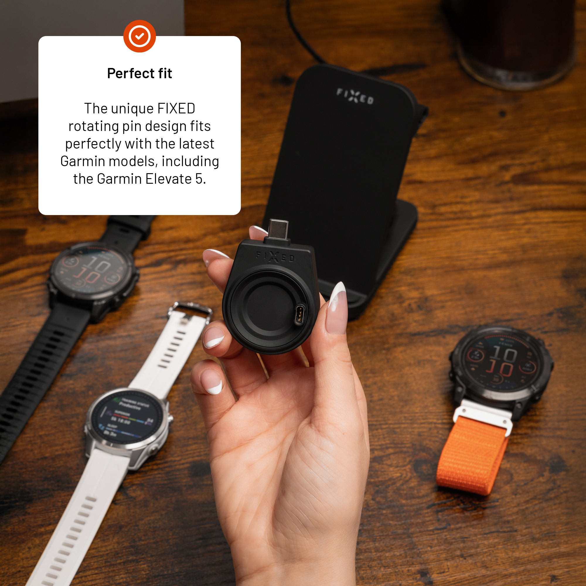 Eine Hand hält einen kleinen, schwarzen FIXED-Ladeadapter, umgeben von Smartwatches auf einem Holztisch.