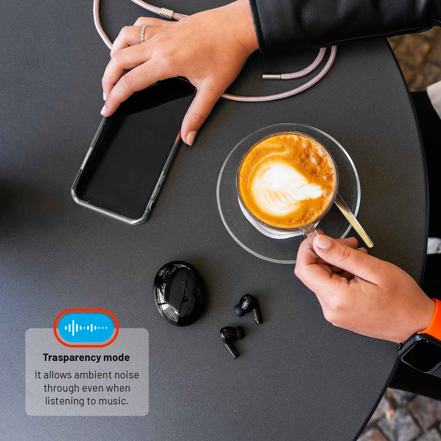 Eine Hand hält ein Smartphone, die andere Kaffee. Auf dem Tisch liegen schwarze Kopfhörer mit Ladecase.